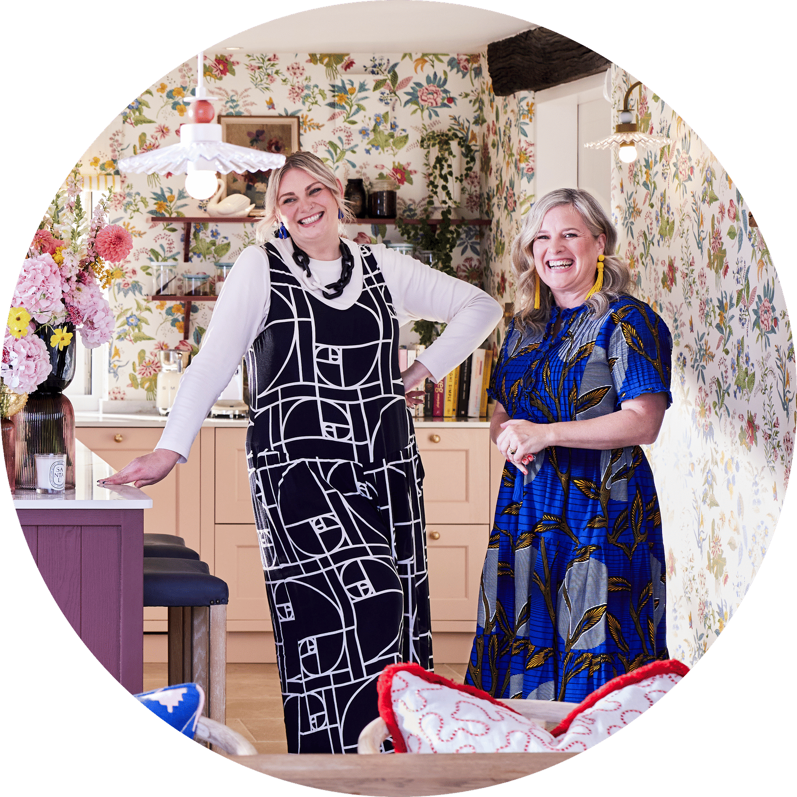 Full room view of Jen from Magnet and Sophie Robinson laughing standing next to the kitchen island in the traditional Shaker style Ludlow kitchen in colours Burlington red, Harvest and Chalk blush matched with a maximalist floral wallpaper.