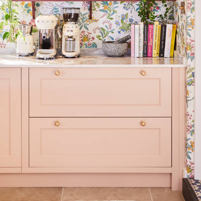 Coffee area in Sophie Robinson's maximalist kitchen, with shaker style Ludlow Chalk blush cabinetry, open shelving on a wall with a floral wallpaper and Smeg coffee machines on countertop.