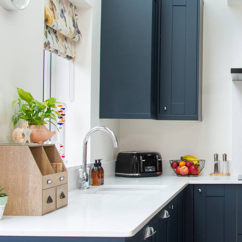 A corner of dark blue shaker kitchen with white worktops, integrated whitegoods and a kitchen island with a breakfast bar.
