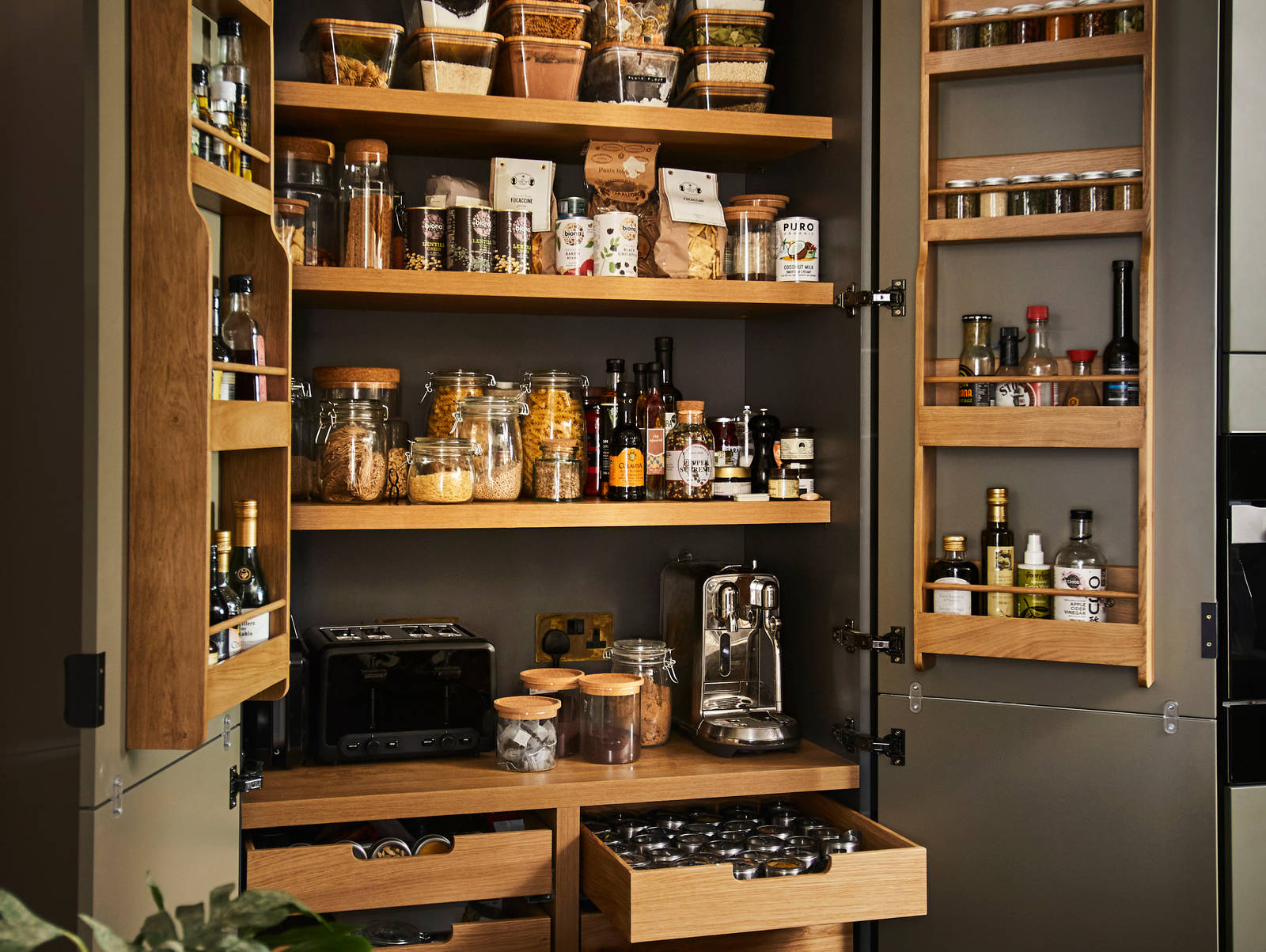 Diagonal view from kitchen island of opened pantry larder with oak interior full of glass jars containing dry goods, a toaster and spices