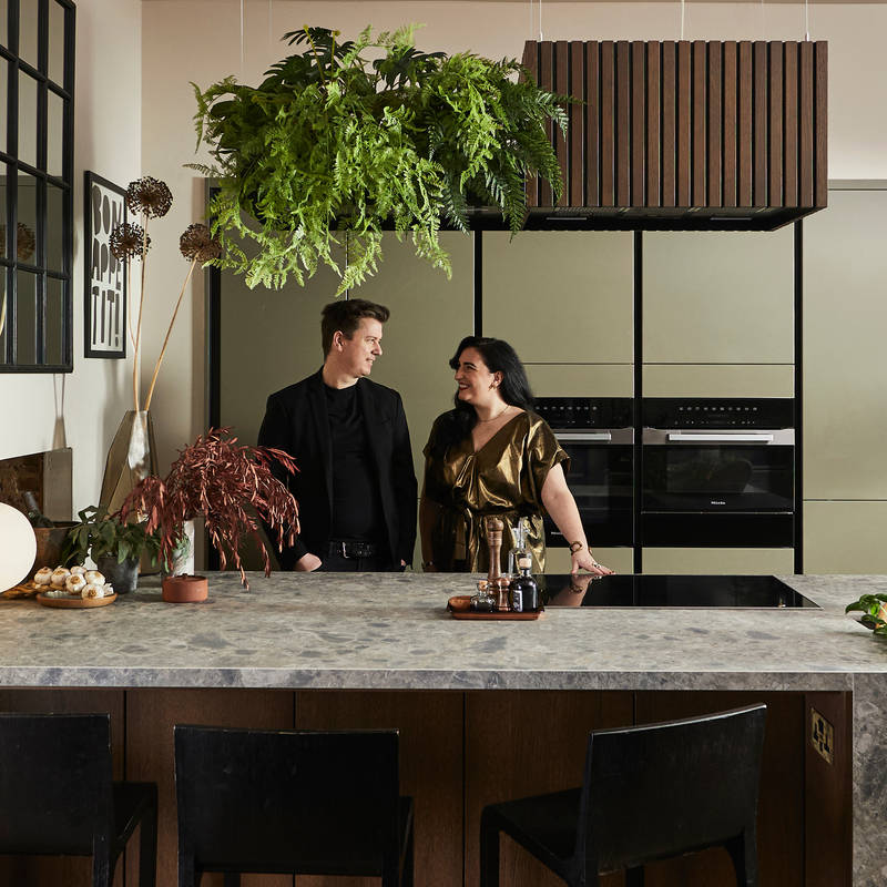 View through opened dark wood doors of the biophilic Dramatic neutral Balmoral Soho and Nordic Nature kitchen from Magnet where James and Ally Dowsing-Reynolds are talking by the kitchen island.