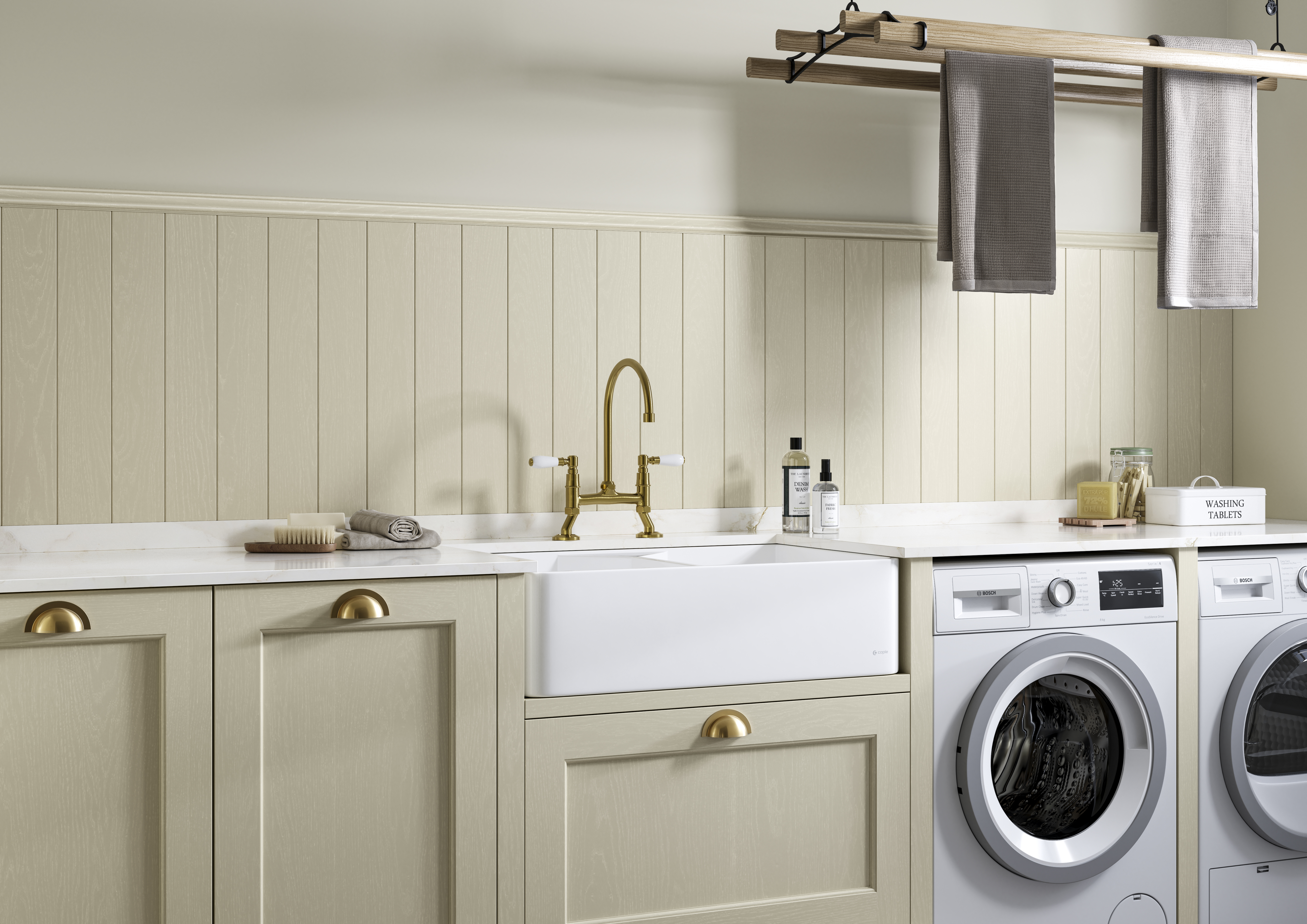 Traditional Shaker-style Ludlow Parchment and French Clay utility room with white ceramic sink, washing machine and tumble dryer with white marbled Silestone worktop and upstand, wallpanel matching the cabinetry, traditional brass tap and towels hanging on wood drying rack.