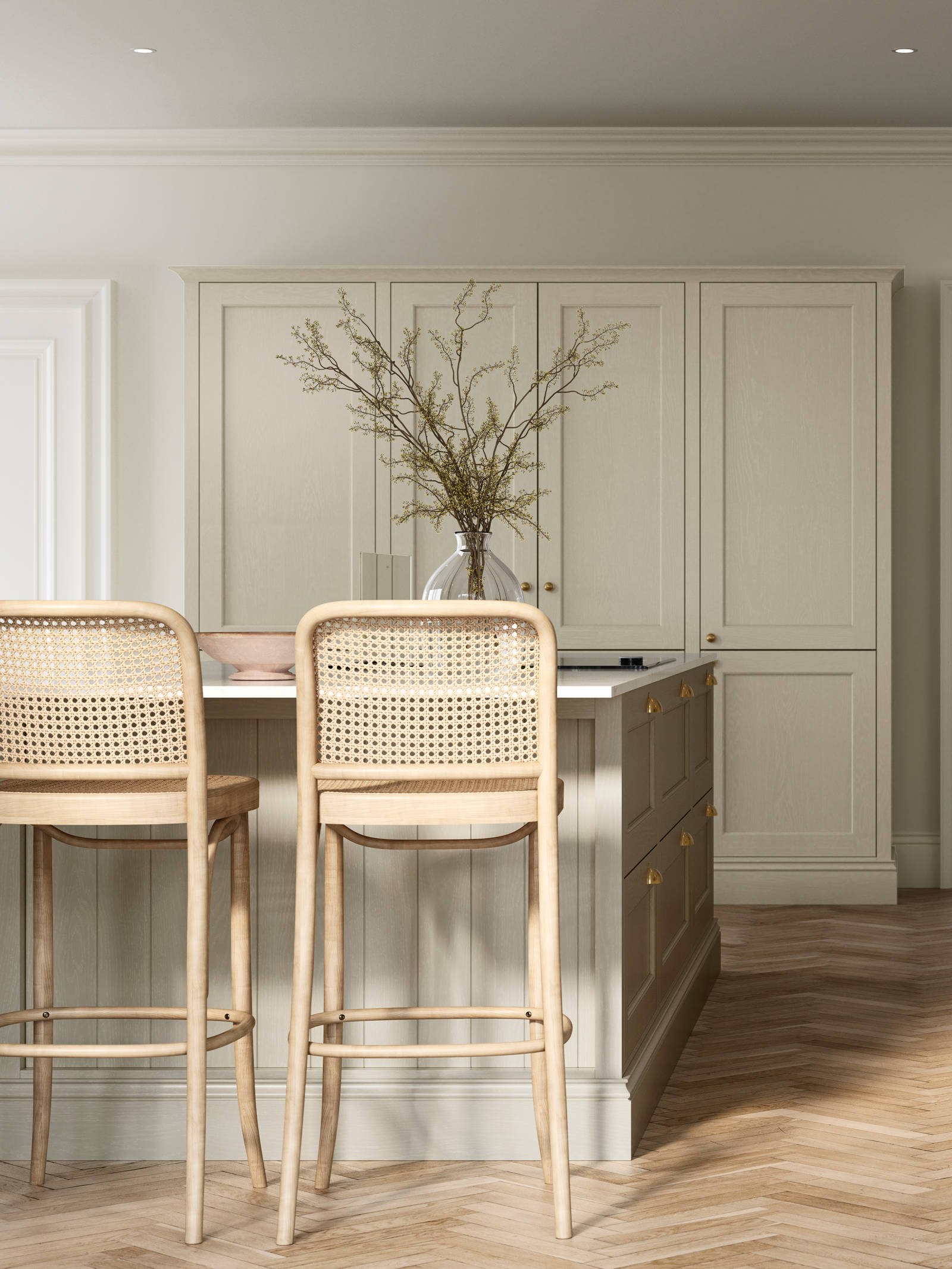 Traditional Shaker-style kitchen Ludlow Parchment with tactile woodgrain, tall cabinets, brass cup handles and knobs, integrated ovens, a kitchen island with rattan bar stools, a vase holding foliage, and light wood herringbone flooring.