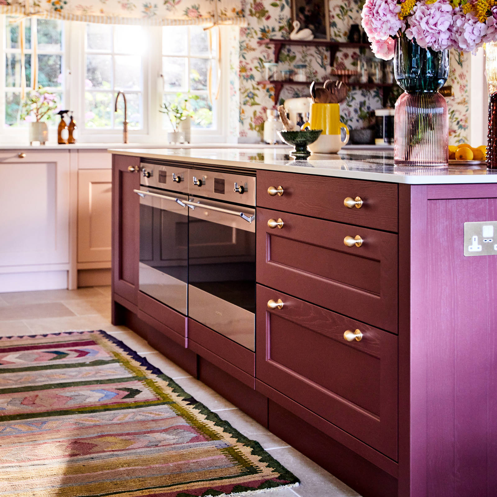 View of Ludlow kitchen island in Burlington Red, ovens in kitchen island and a woven colourful rug on the tile floor.