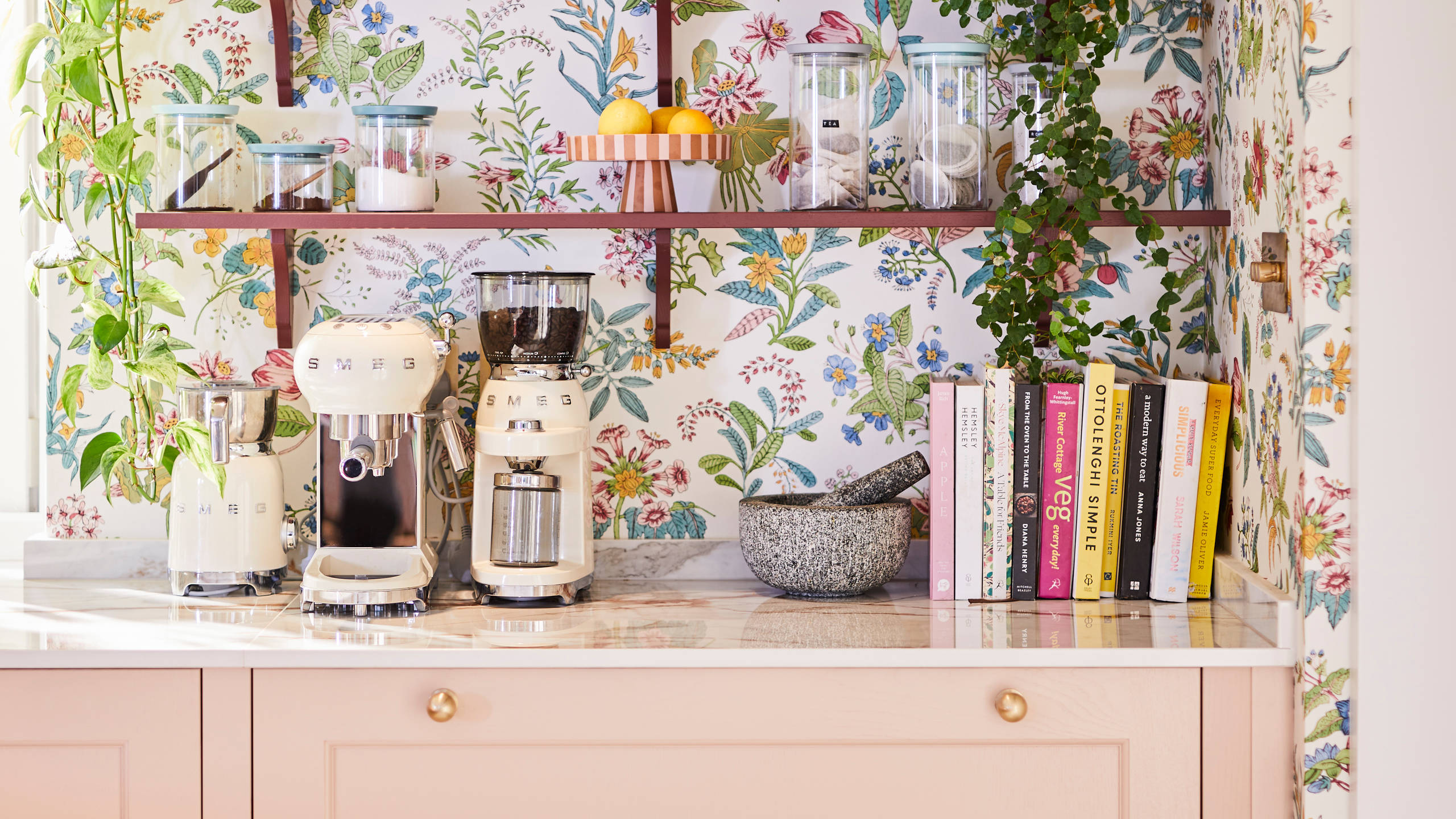 Coffee area in Sophie Robinson's maximalist kitchen, with shaker style Ludlow Chalk blush cabinetry, open shelving on a wall with a floral wallpaper and Smeg coffee machines on countertop.