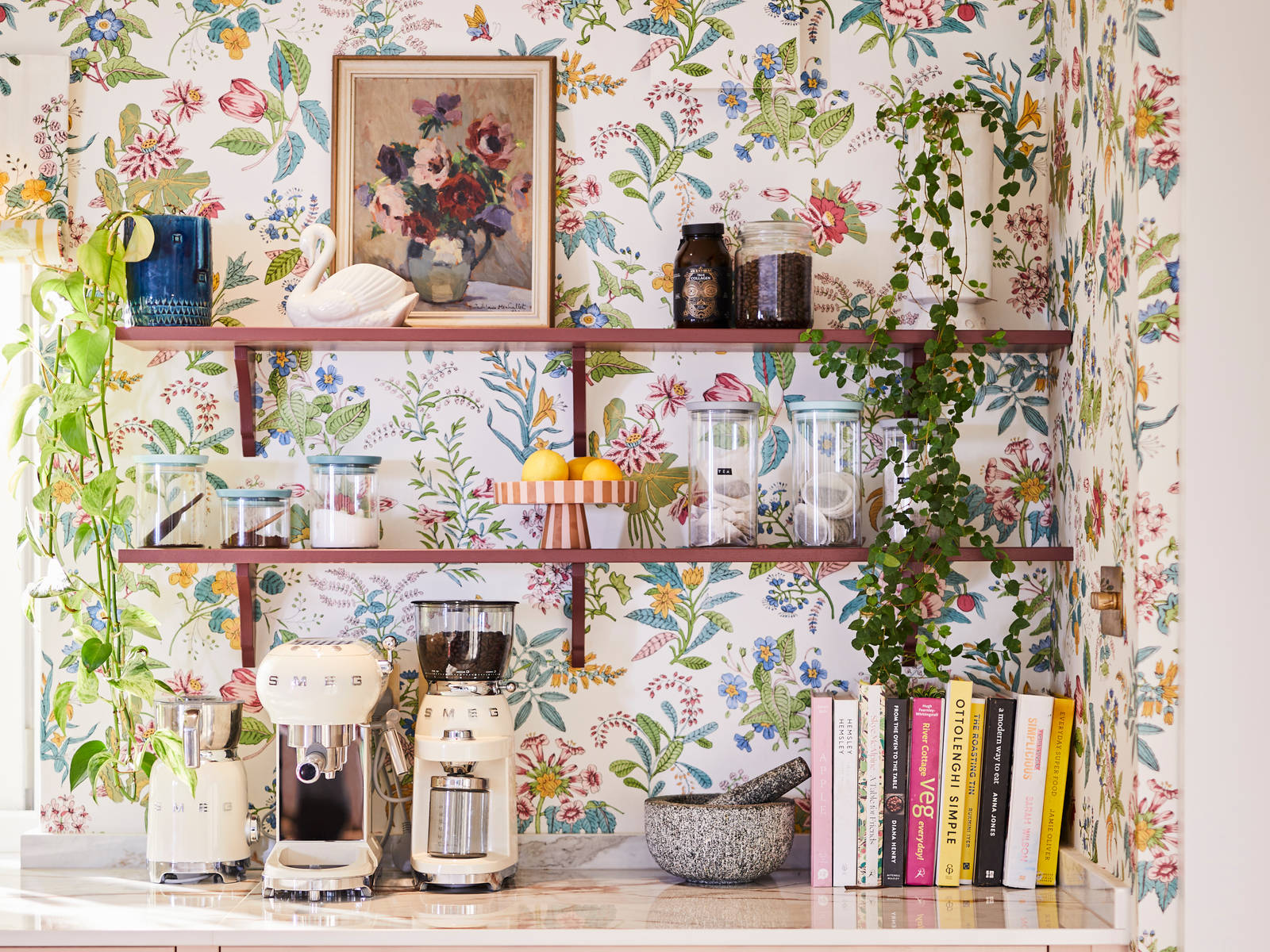Coffee area in Sophie Robinson's maximalist kitchen, with shaker style Ludlow Chalk blush cabinetry, open shelving on a wall with a floral wallpaper and Smeg coffee machines on countertop.
