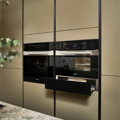 Built-in Ovens with opened warming drawer in modern slab front Balmoral Soho cabinetry from Magnet, Dekton in Ceppo worktop of kitchen island in foreground.
