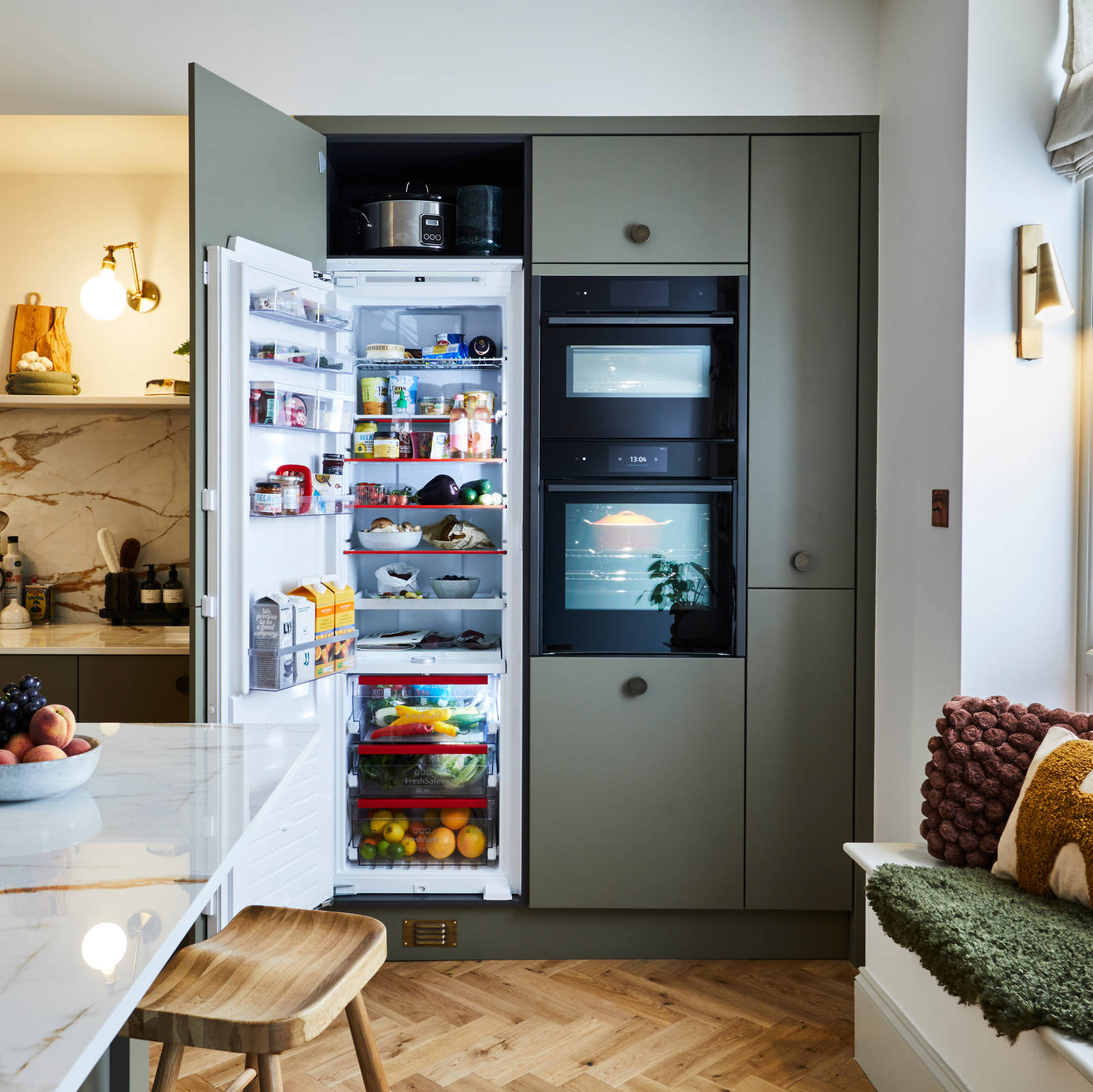Open fridge in Soho Balmoral tall cabinets with integrated ovens and a bench in the window, next to a wall of open wood shelves decorated with plants, candles and wine glasses.