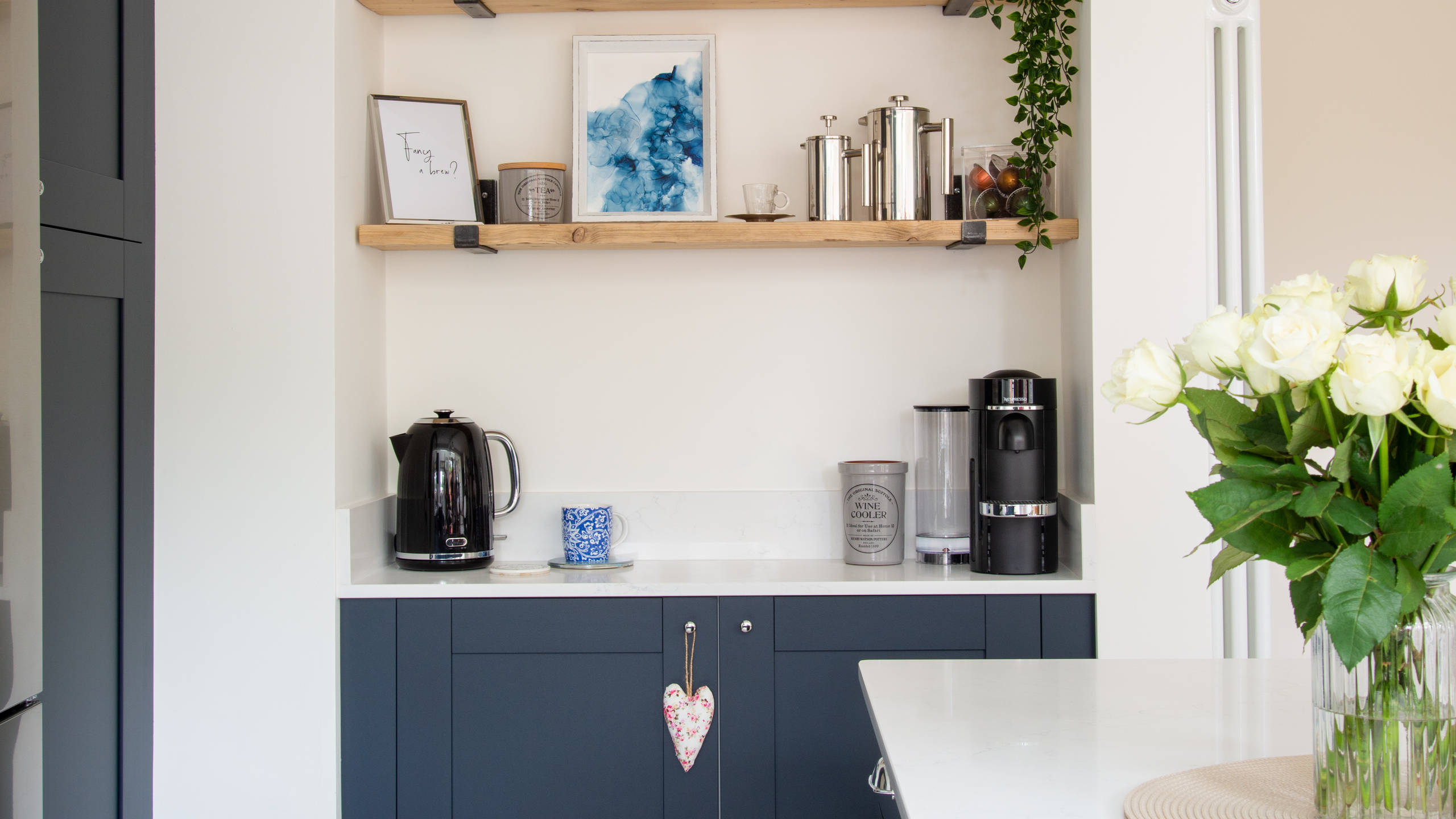Dark blue shaker cabinets, white worktop, kettel and coffee machine with open wooden shelves on wall above.