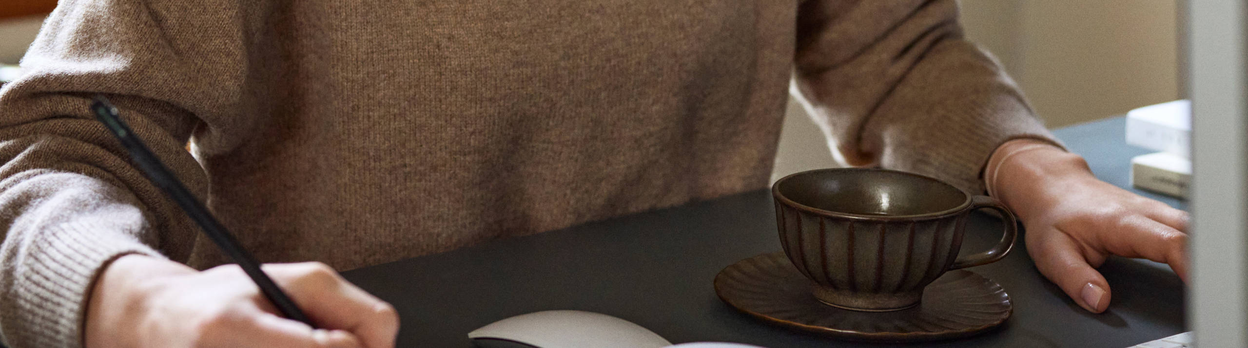 Ung kivnna i naturfärgad stickad tröja antecknar i ett skrivblock framför datorn