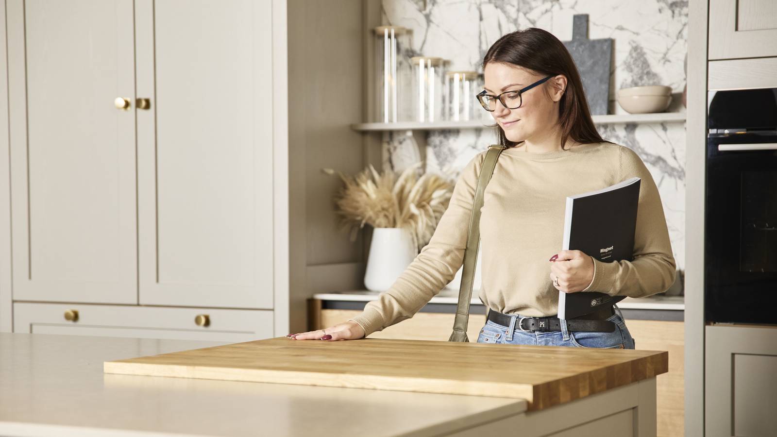 Woman holding a brochure feeling the wood worktop with her hand in a neutral shade Shaker style kitchen in a Magnet Showroom