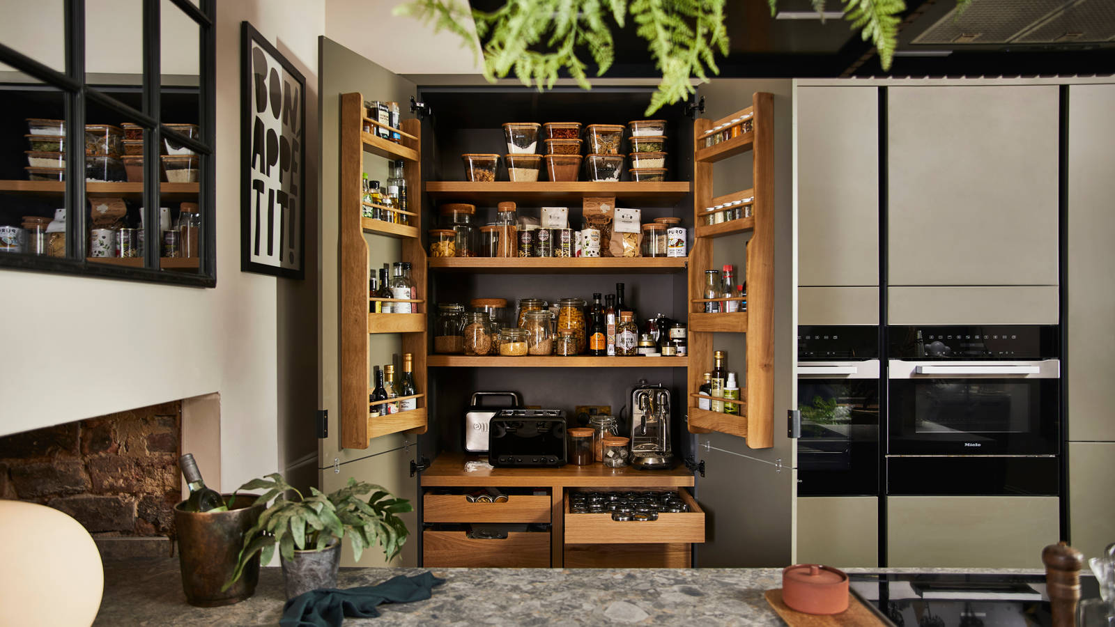 View from kitchen island of opened pantry larder with oak interior full of glass jars containing dry goods, a toaster and spices