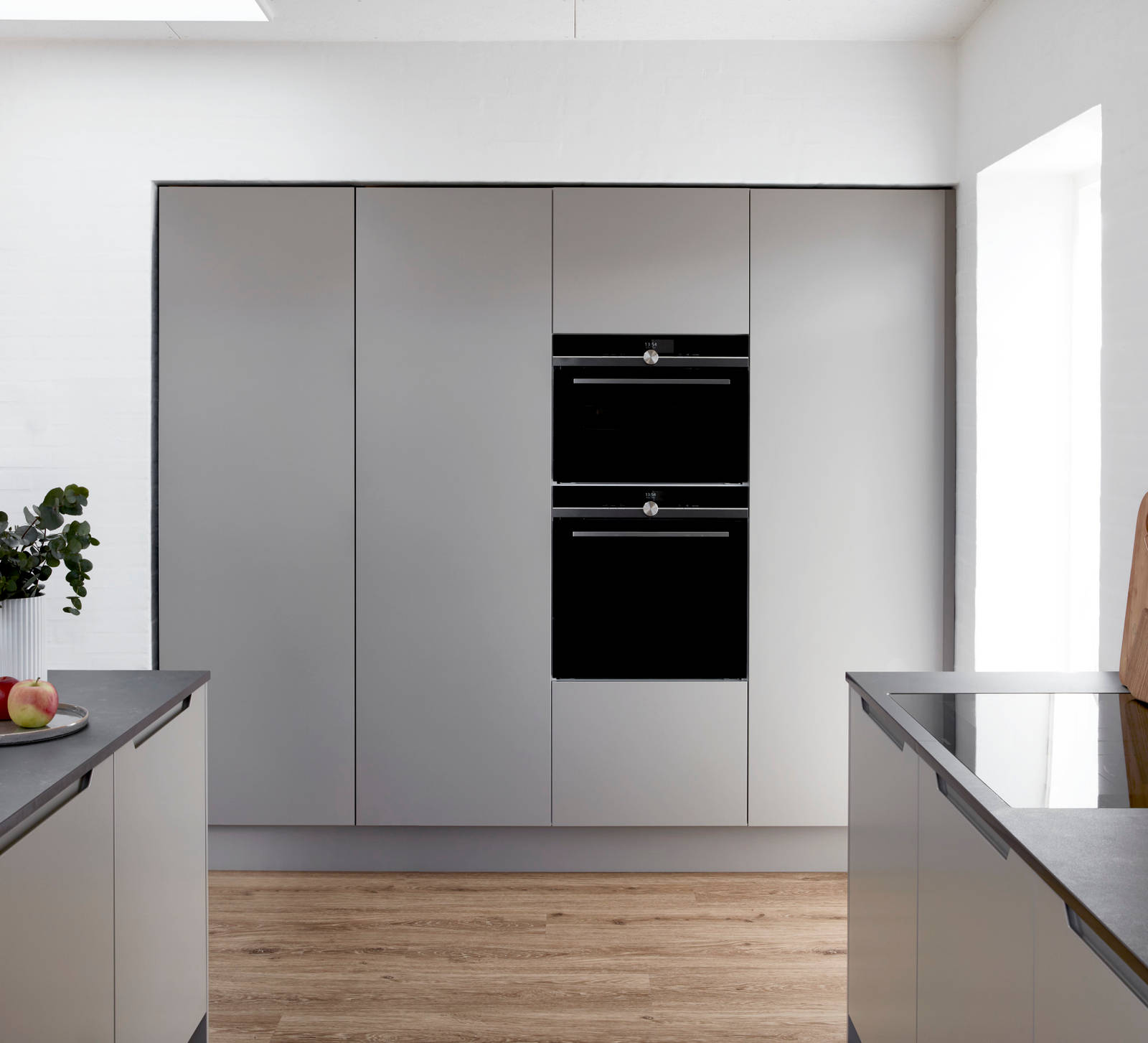 Højskabe med indbygningsovne i grå køkken Urban Søsten med køkkenø fra INVITA med indfræsede greb og sort bordplade