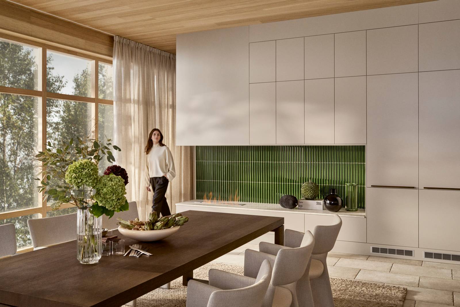 Kvinde kommer ind i rummet med spisebord i tre, benk og toppskabe Athena Sand med grønne fliser bak