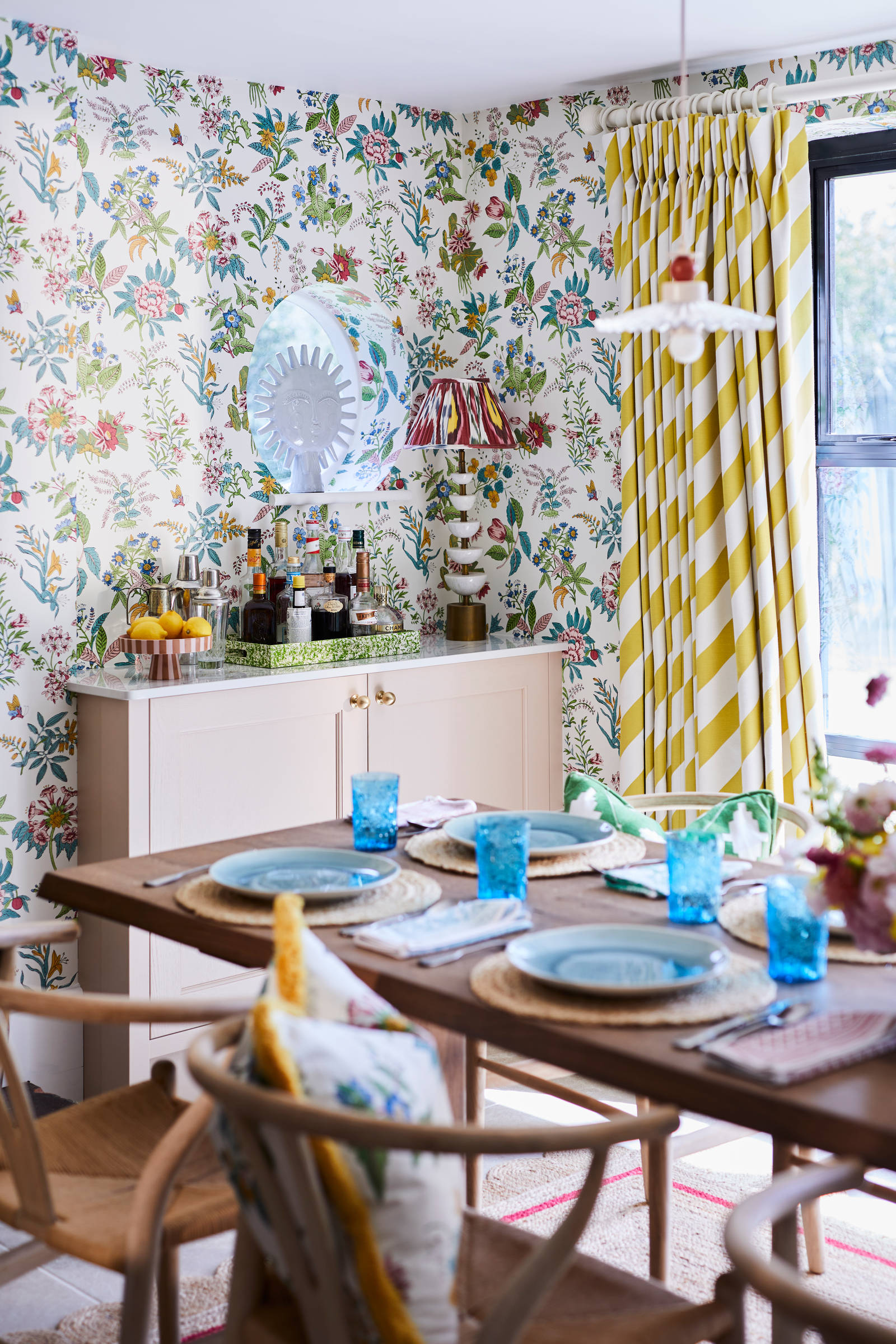 Cocktail corner in Sophie Robinsons maximalist Ludlow kitchen from Magnet, with a dining room table in foreground, floral pattern wallpaper and striped curtains.
