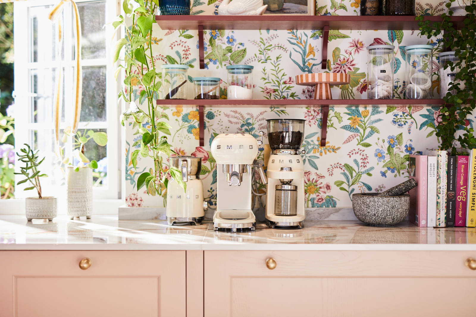 Close-up of Smeg coffee machines on counter top in the coffee area in Sophie Robinson's maximalist kitchen, with shaker style Ludlow Chalk blush cabinetry, open shelving on a wall with a floral wallpaper.