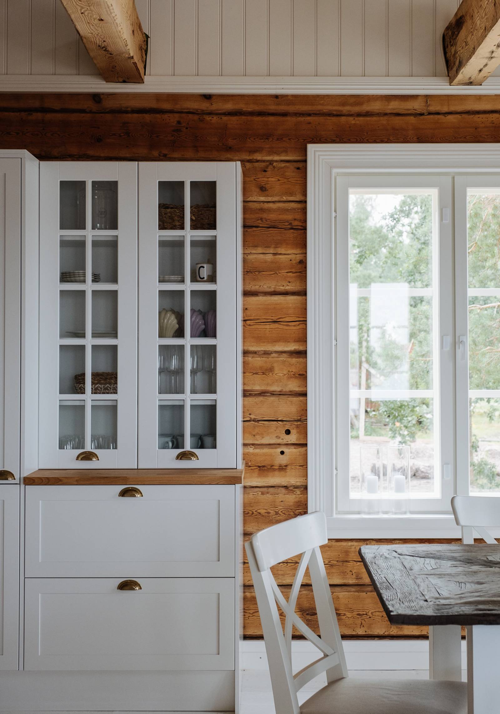 Keittiö, jossa karmiovet ja yhteensopivat maalatut vitriinekaapit 1800-luvun hirsitalossa, tammitasot ja messinkiset kupinkahvat.