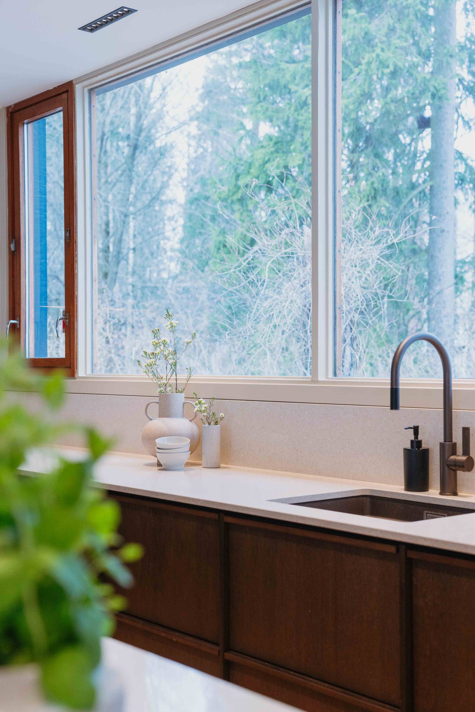 Svart blandare och diskho framför stora fönster i Liv och Hallvards Signatur Ekbacken-kök från Marbodal, med brunbetsad fanerad eklucka, matchande handtag i trä och bänkskiva i kvartskomposit