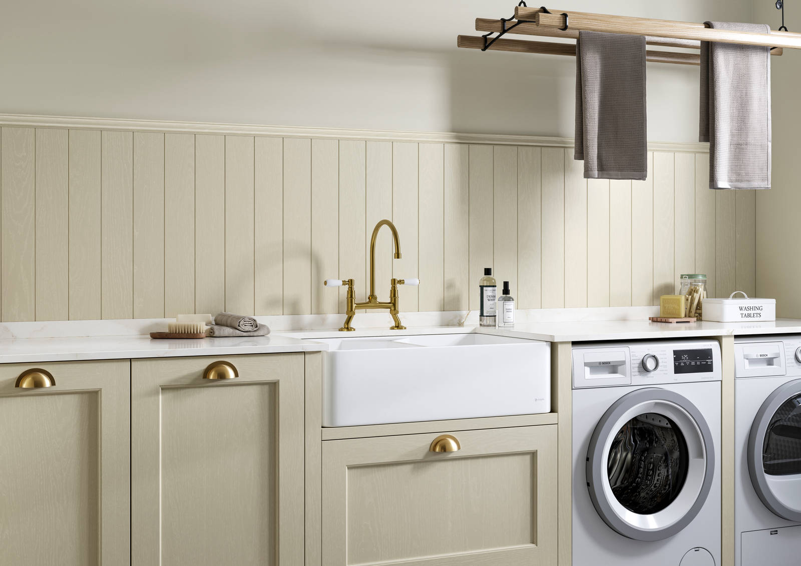 Traditional Shaker-style Ludlow Parchment and French Clay utility room with white ceramic sink, washing machine and tumble dryer with white marbled Silestone worktop and upstand, wallpanel matching the cabinetry, traditional brass tap and towels hanging on wood drying rack.