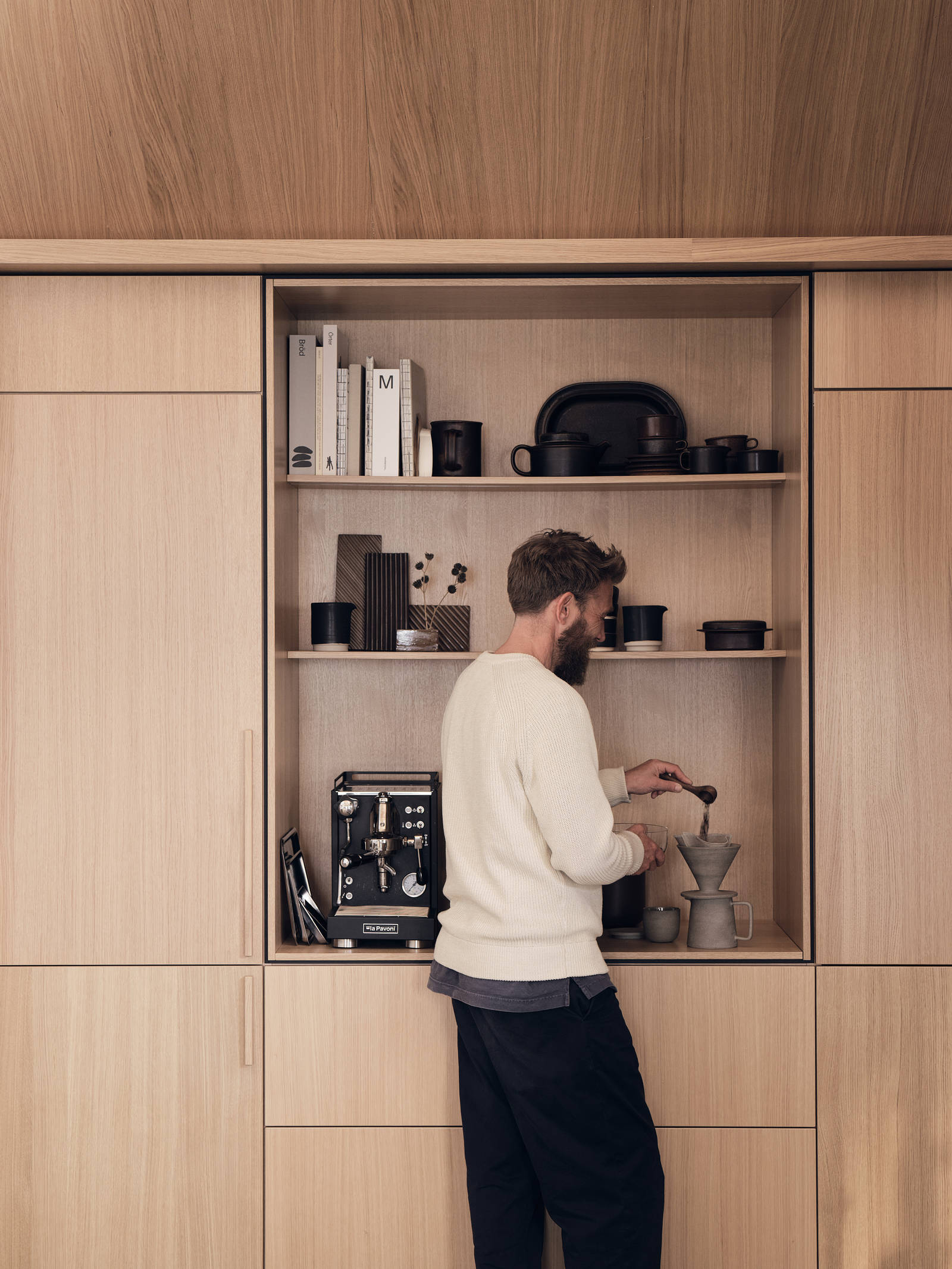 En man mäter upp kaffe i köket Ekbacken natur från Marbodal i ekfanér med vertikal träådring. Högskåp och ett öppet skåp med plats för kaffemaskin, inramad med en skuggning vilket skapar en arkitektonisk premiumkänsla.
