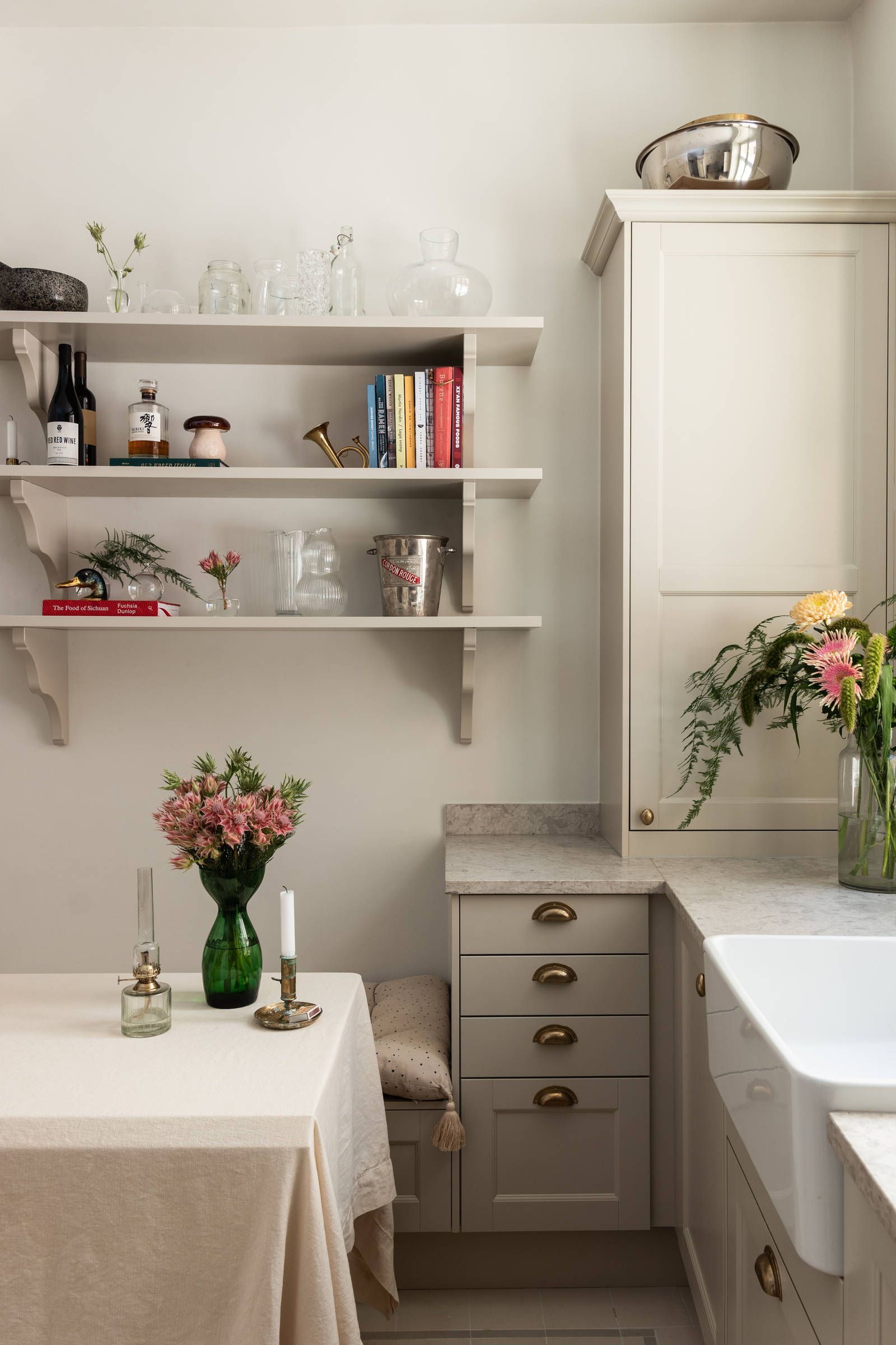 Sandra Beijers kök från Marbodal, lantliga luckan Fagerö i beige kulören Sand, med bronsfärgade handtag, diskho i vitt porslin, öppna vägghyllor i matchande beige kulör, en vit duk och mässingsljusstakar på matbordet.