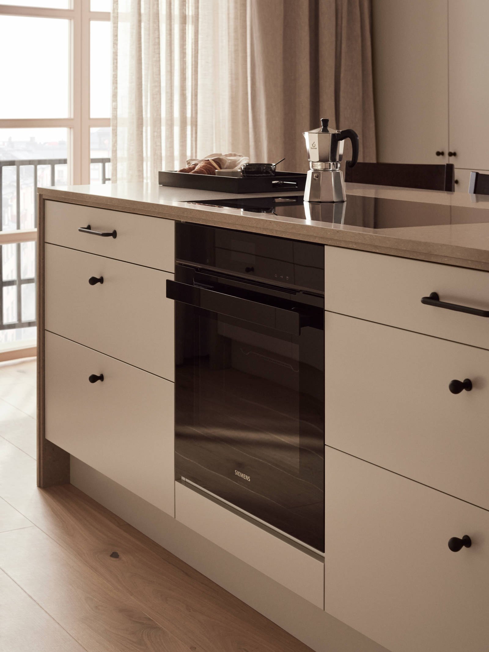 Inbyggnadsugn och induktionshäll från Siemens i köksö i köket med beige slät kökslucka Arkitekt Plus gryning, bänkskiva och dekorativ kant i kompositsten, svarta knoppar