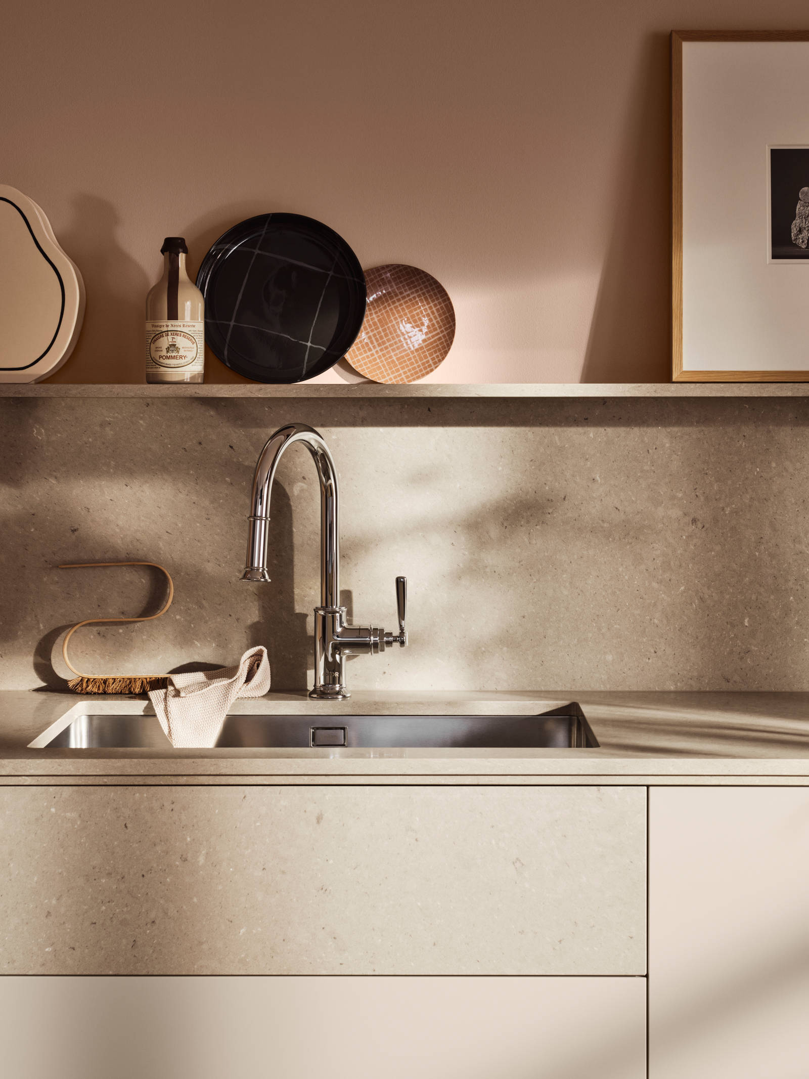 Köksbänk med beige slät kökslucka Arkitekt Plus gryning, bänkskiva, stänkskydd och vägghylla i matchande kompositsten, dekorationer på hyllan, falsk belfast-diskho i matchande dekorativ stenfront.