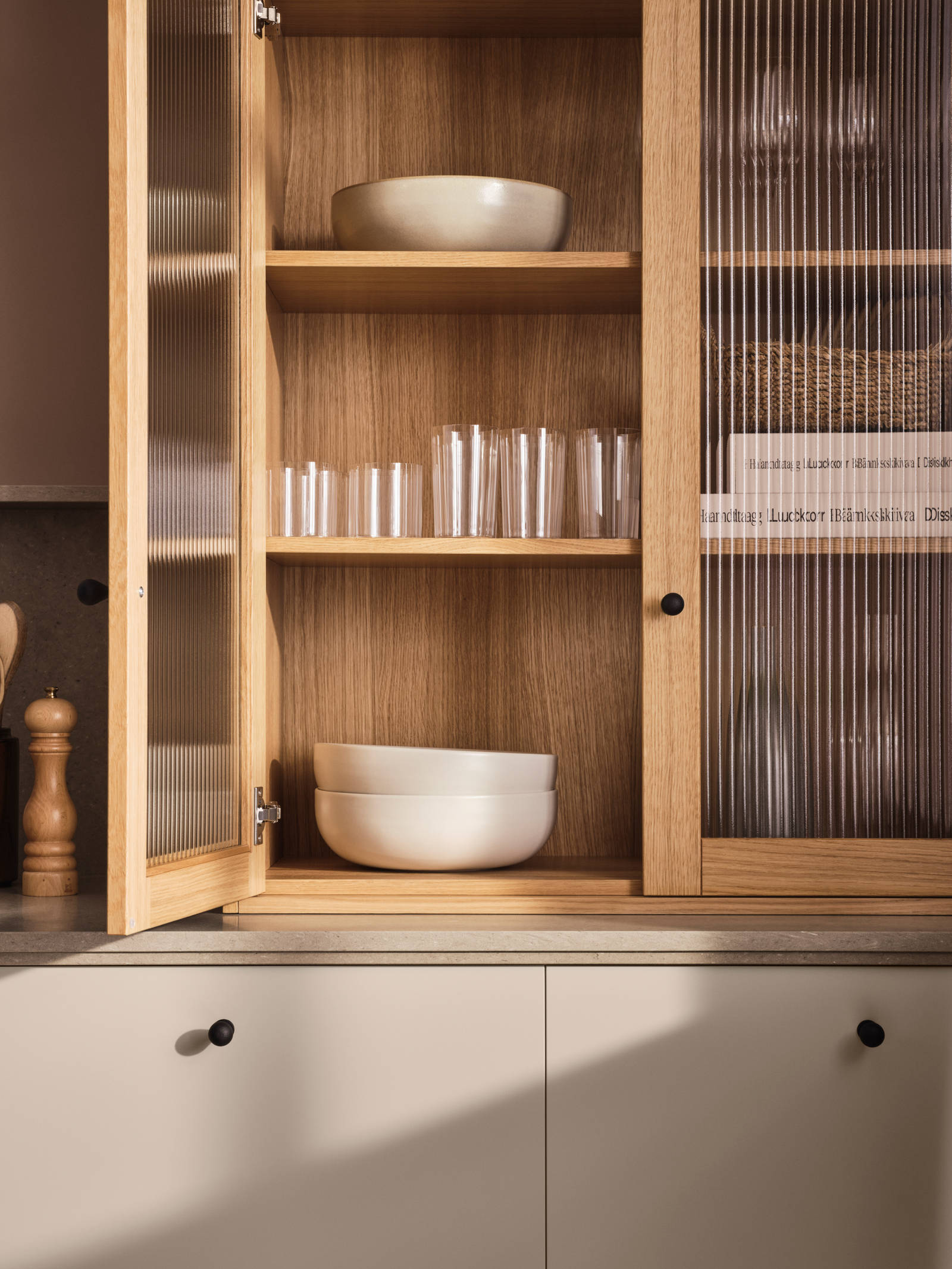 Vitrinskåp ovanpå köksbänk i beige kök Arkitekt plus gryning från Marbodal, öppen lucka i vitrinskåp med träram i natur ek och räfflat glas, keramik och dricksglas