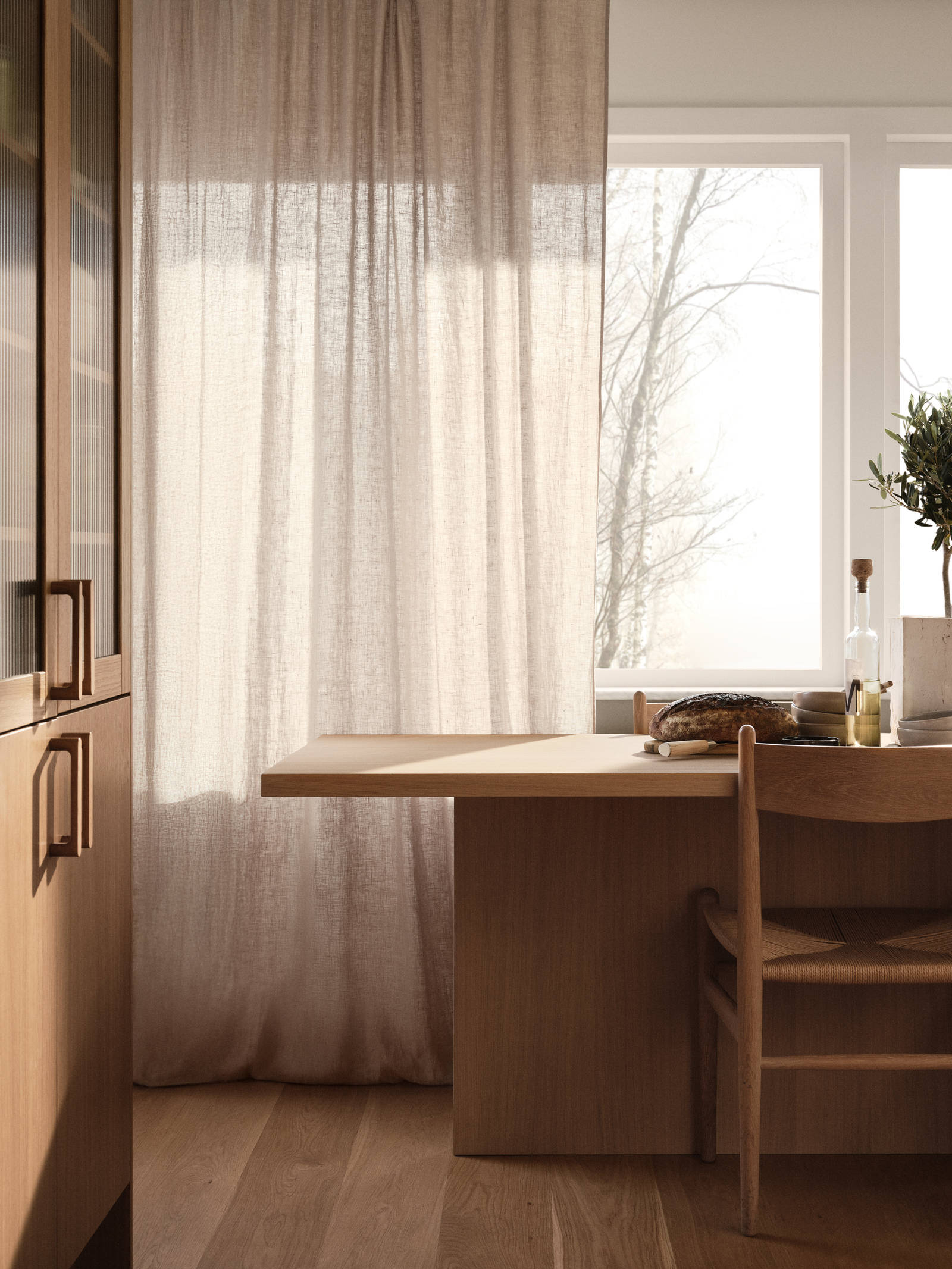 Ett ljust rum med stora fönster, ljusa lingardiner, bord och stol i ljust trä, högskåp med eklucka och vitrinluckor med ram i ek hemma hos Siri Barje.