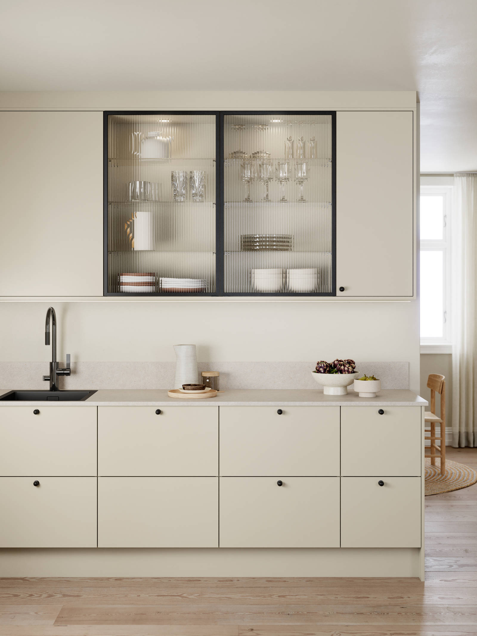 Beige melaminkök med svarta knoppar, svart blandare och vitrinlucka med svart aluiminiumram och räfflat glas.