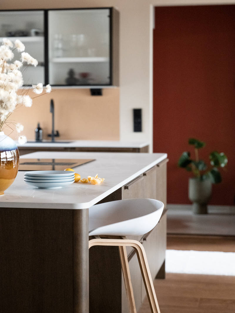 Keittiösaareke pyöristetyllä kulmalla, kaapit laatikoilla ja istumatilaa valkoisella ja puisella baarijakkaralla