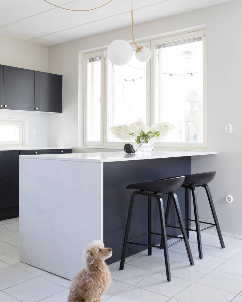 Perhekoira lattialla tummansinisessä Meri maalatussa sileässä keittiössä, jossa valkoinen marmorinen työtaso ja siihen sopiva splashback, keittiösaareke istuinalueella, messinkinupit ja messinkihana