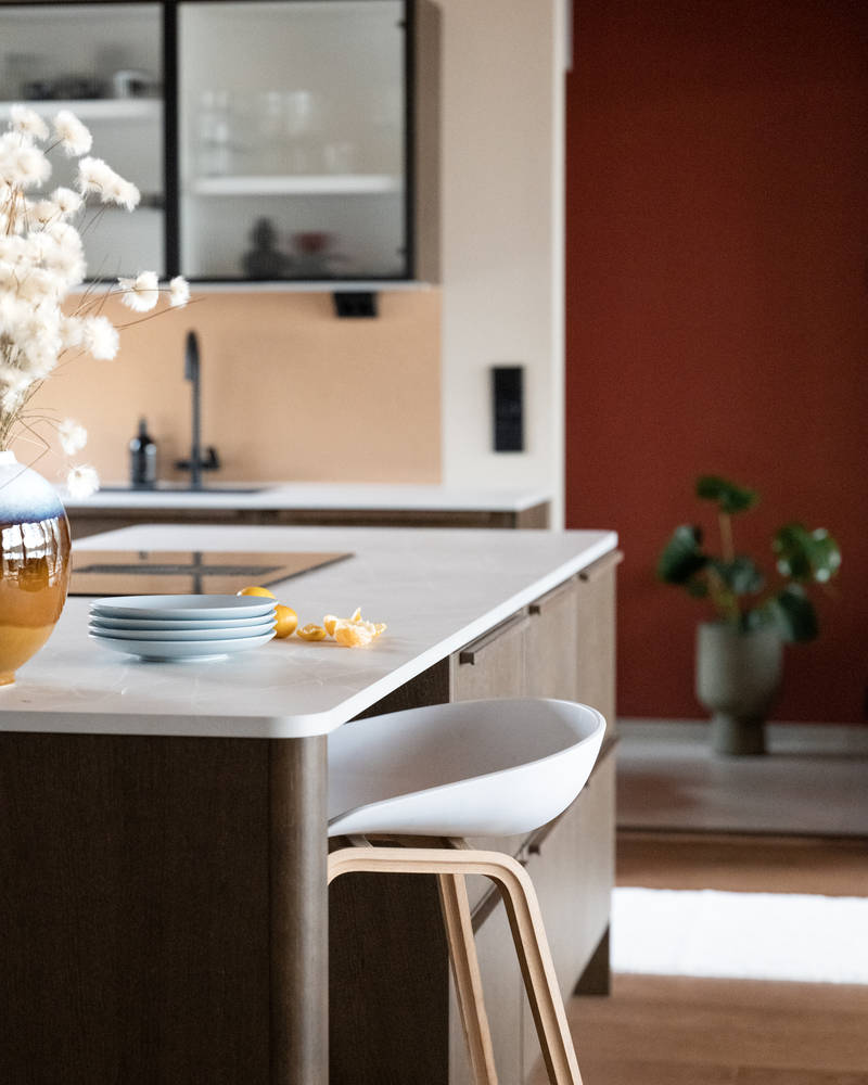 Keittiösaareke pyöristetyllä kulmalla, kaapit laatikoilla ja istumatilaa valkoisella ja puisella baarijakkaralla