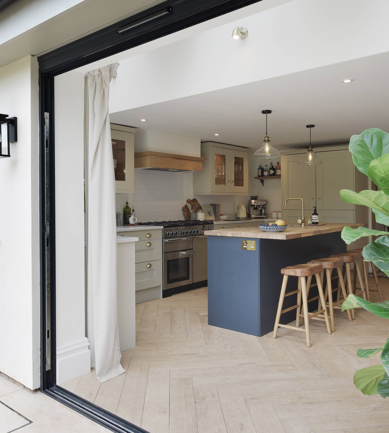 View of the country cottage throught opened patio doors of the Shaker-style kitchen in shade Sage with a midnight blue kitchen island.
