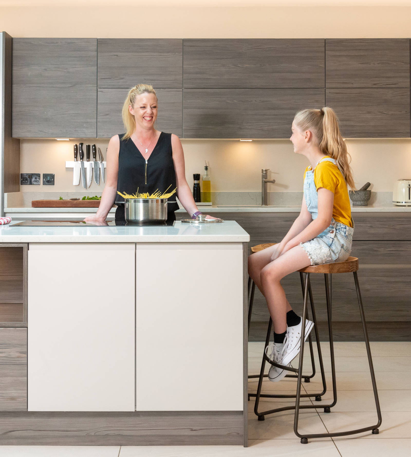 Magnet kitchen customer Caroline boiling pasta and smiling to her daughter sitting on a barstool at the kitchen island