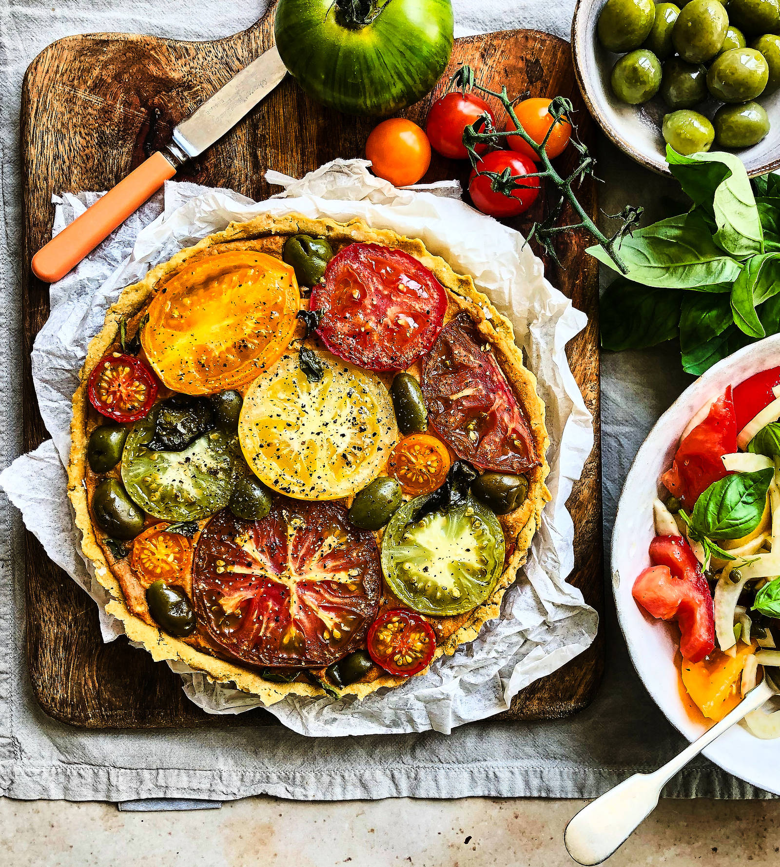 Tomato and olive tart. served on a rustic wooden board, with a handmade ceramic bowl of olives next to it, and a salad bowl
