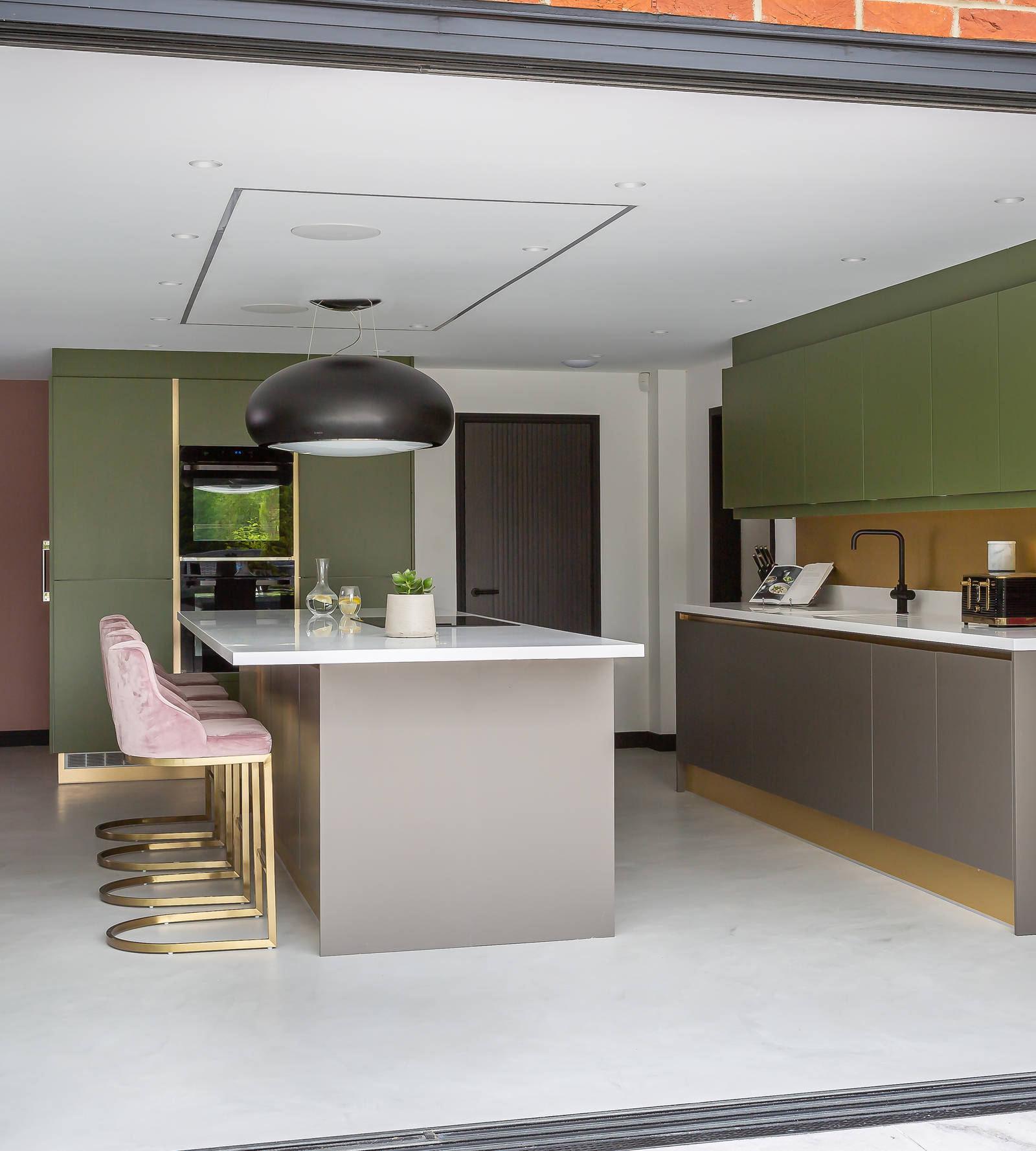 Goddens family kitchen - handleless in grey and green shades, with white worktop and a kitchen island with breakfast bar, brass details and brass chairs.