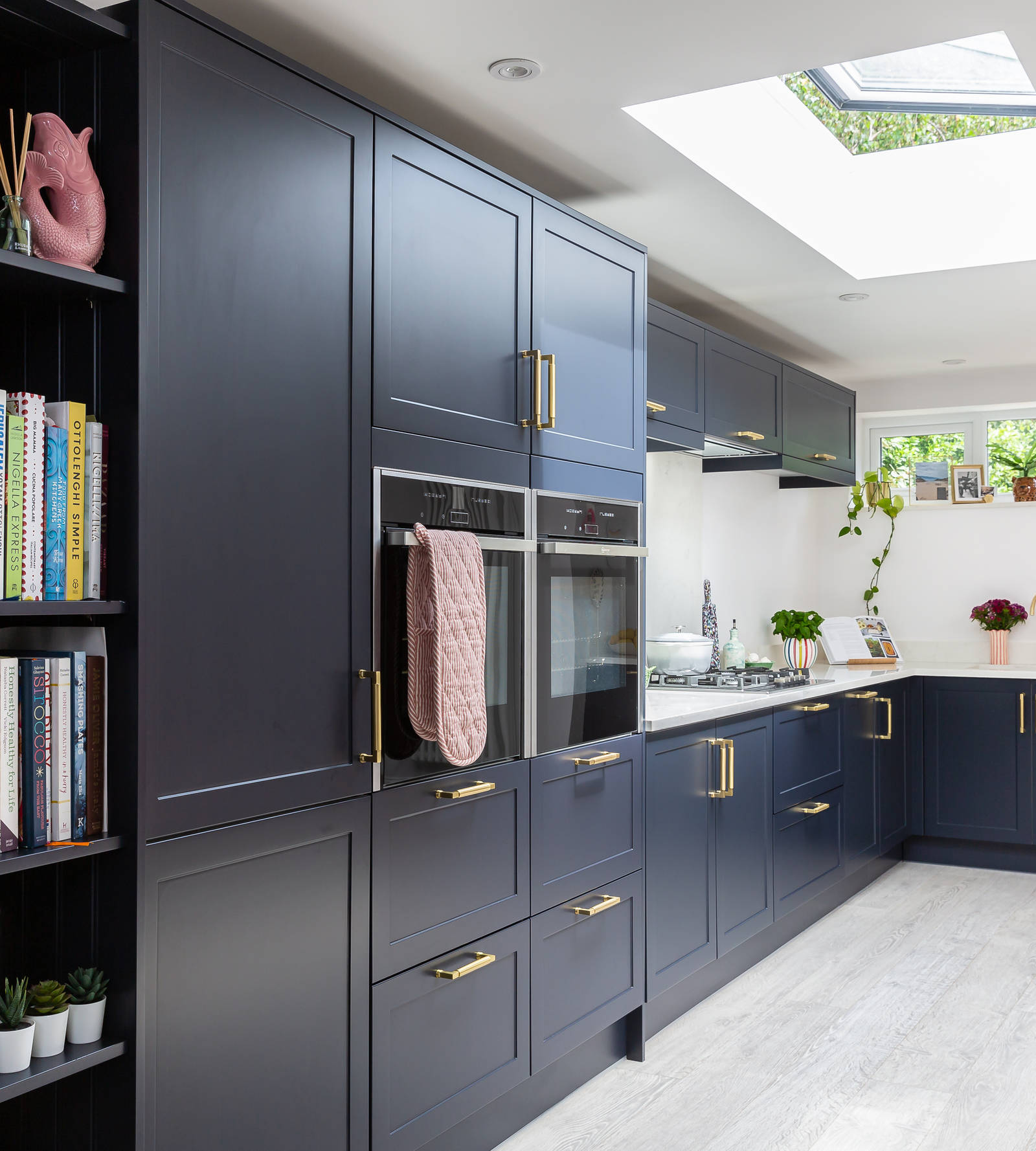 Dark blue shaker kitchen with white worktop, integrated ovens in tall cabinets and open shelves woth cookbooks