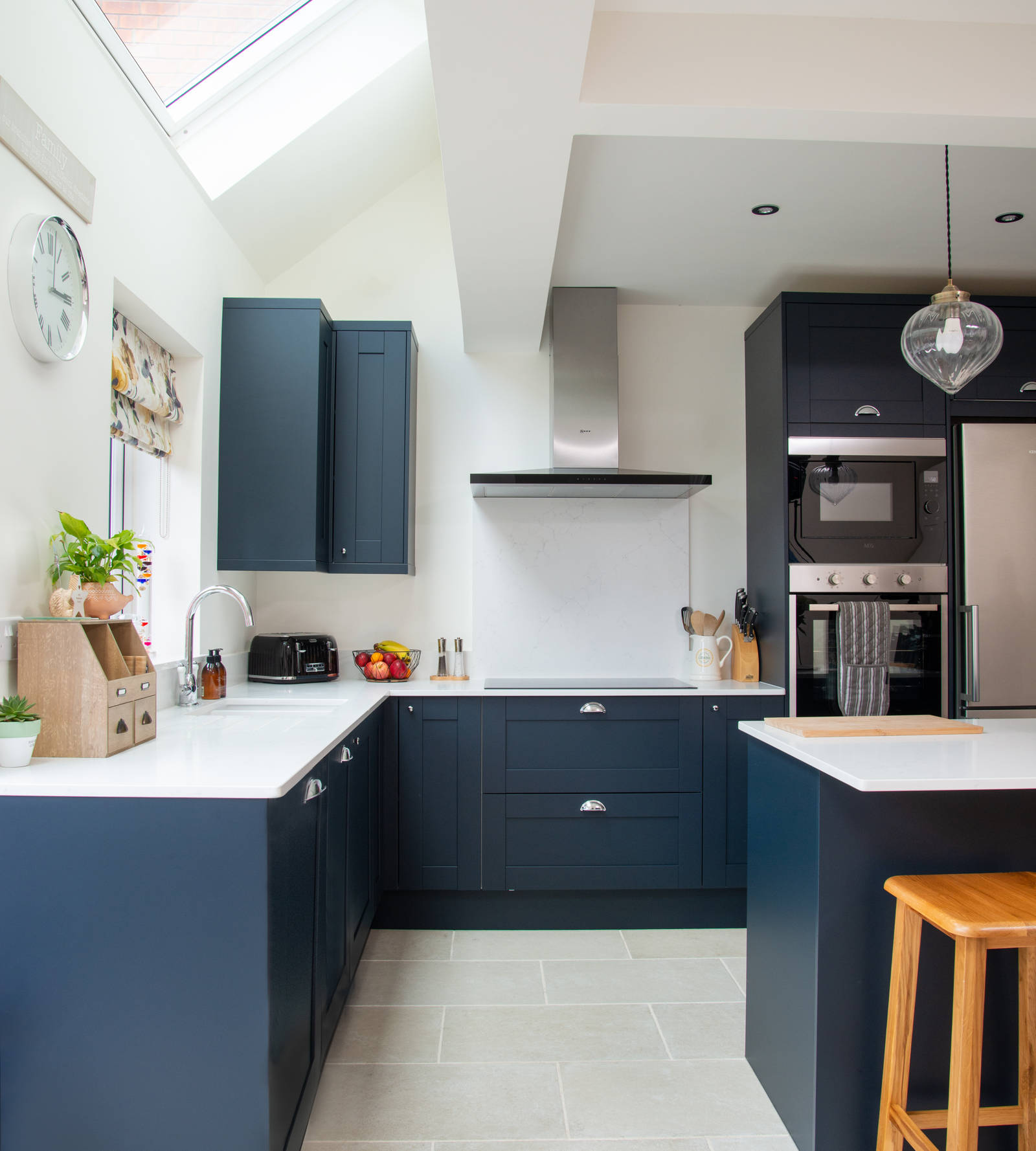 A corner of dark blue shaker kitchen with white worktops, integrated whitegoods and a kitchen island with a breakfast bar.