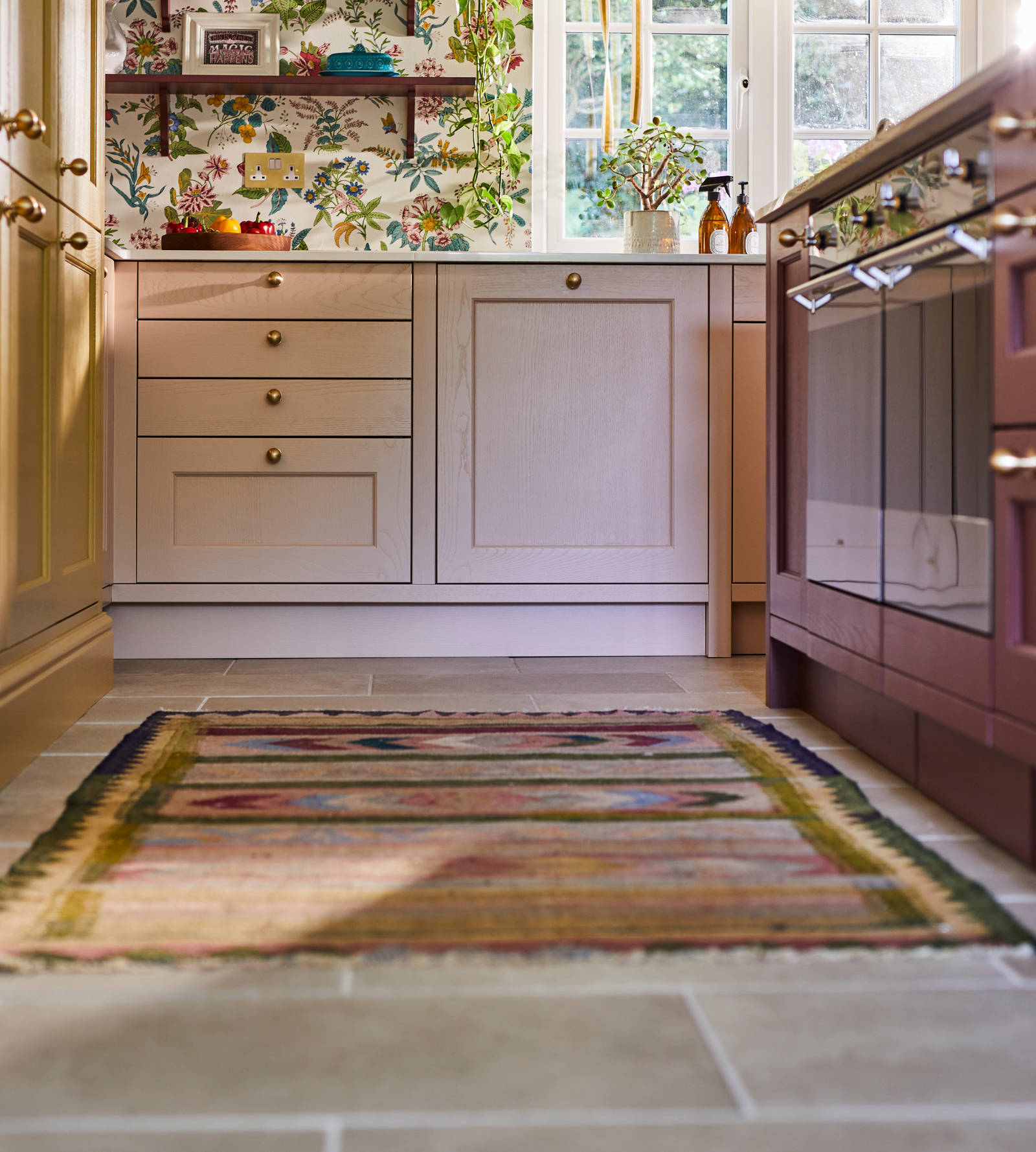 View of Ludlow cabinetry in colours Harvest, Chalk blush and Burlington Red, and a woven colourful rug on the tile floor.