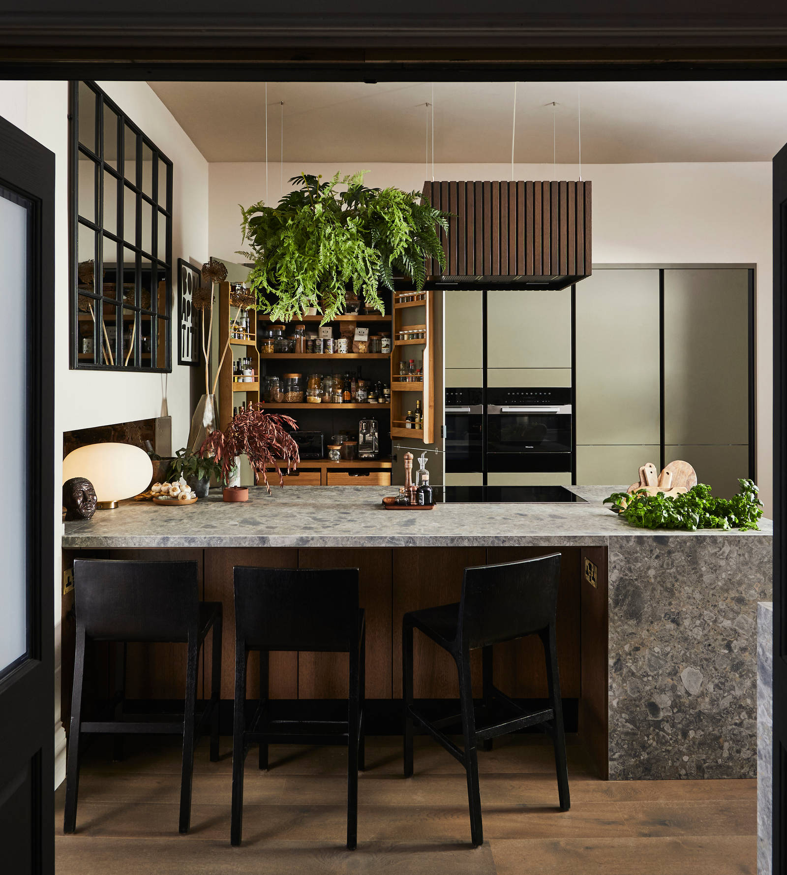 View through opened dark wood doors of the biophilic Dramatic neutral Balmoral Soho and Nordic Nature kitchen from Magnet, with the pantry larder cabinet opened revealing oak interiors.