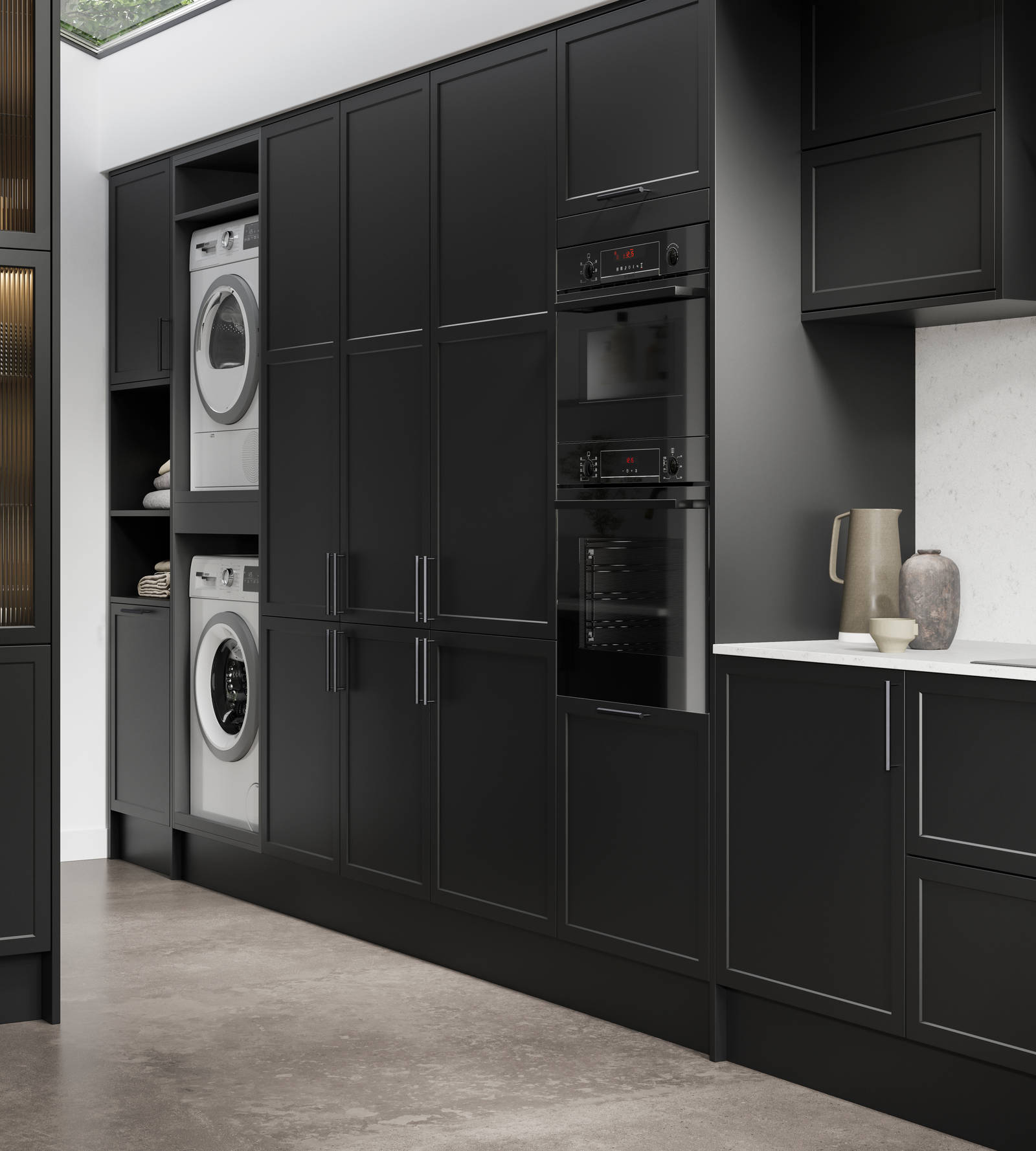 View of Modern Shaker Brunswick Sumi Black laundry room from kitchen, with integrated laundry machine and dryer as well as ovens in tall cabinetry, white worktop and splashback Silestone Miami Vena and vitrine cabinets with fluted glass doors.