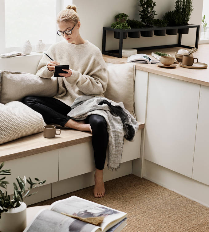 Kvinna med glasögon sitter på bänken i köket Arkitekt Plus Frost från Marbodal, med en filt i knät och anteckar i ett block, med en kaffekopp framför sig.