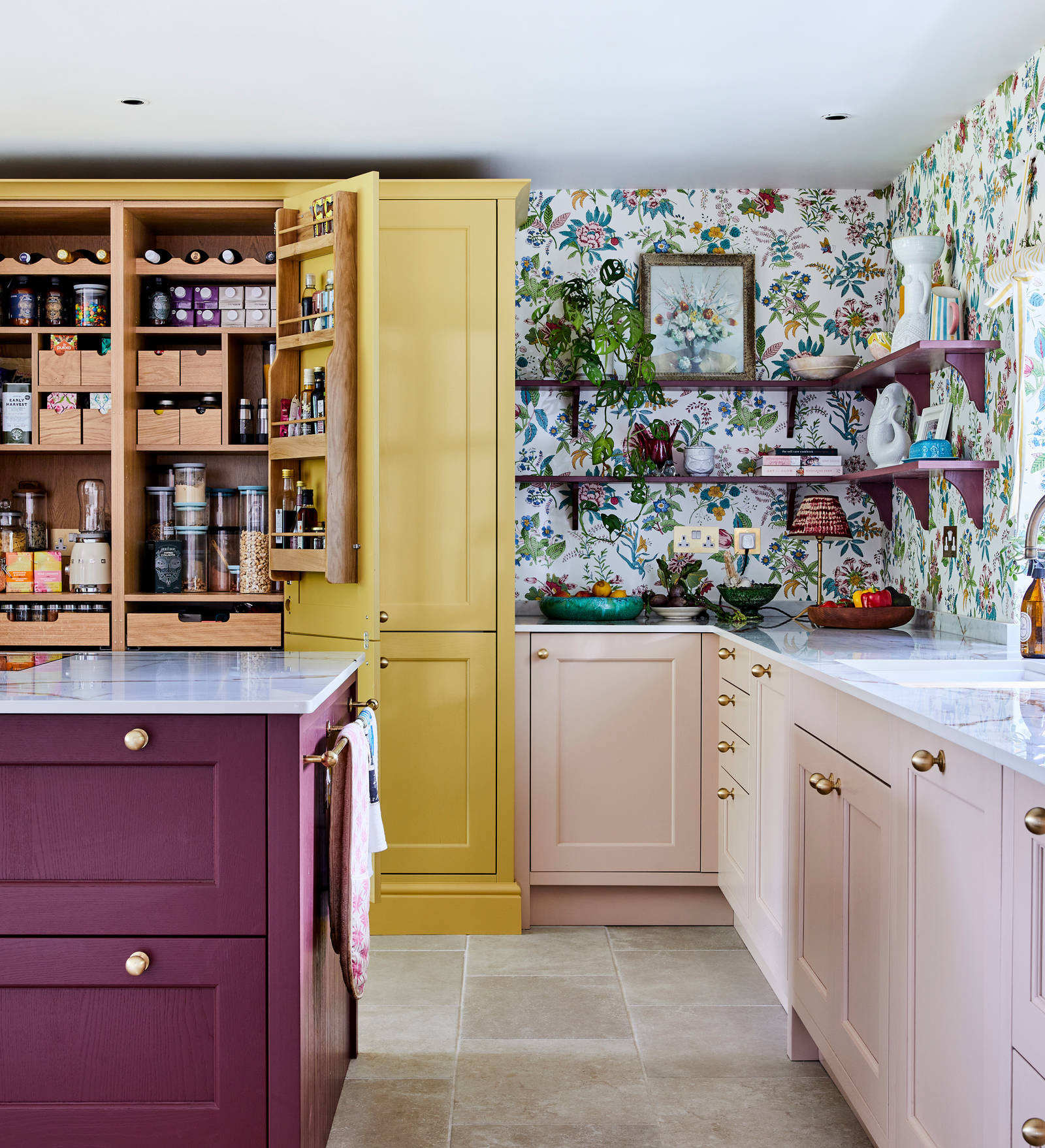 Wide image of sink, kitchen island an opened pantry larder. Ludlow cabinetry from Magnet in colours Burlington Red, Chalk blush and Harvest, wallpaper with a colourful floral pattern.