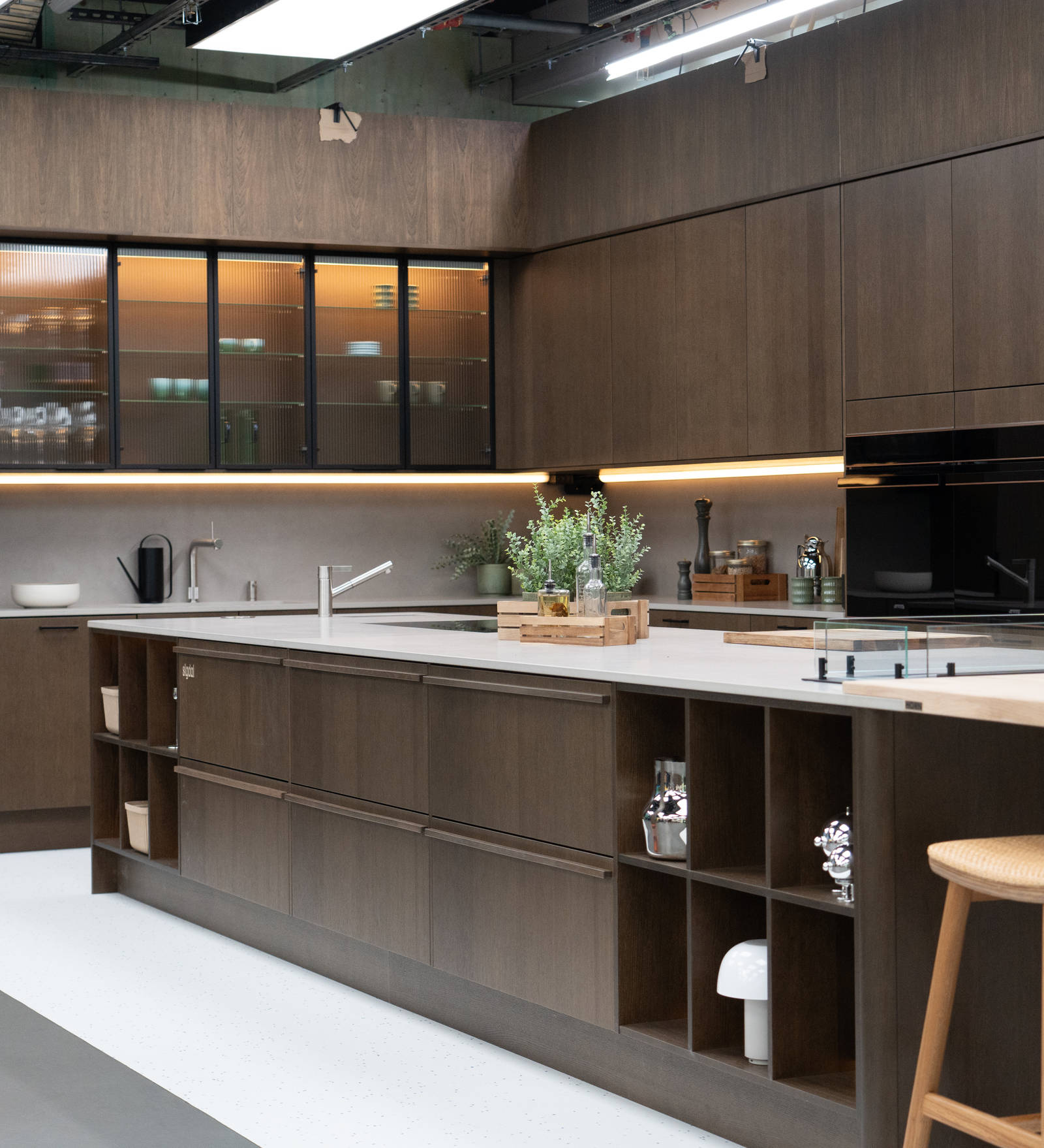 Kjøkken i modell Favn fra Sigdal i lekker eikefinér, som er beiset i fargen Nøtt, kjøkkenøya og vitrinedører med bølget glass og sorte aluminiumsrammer i God Morgen Norge studion sin