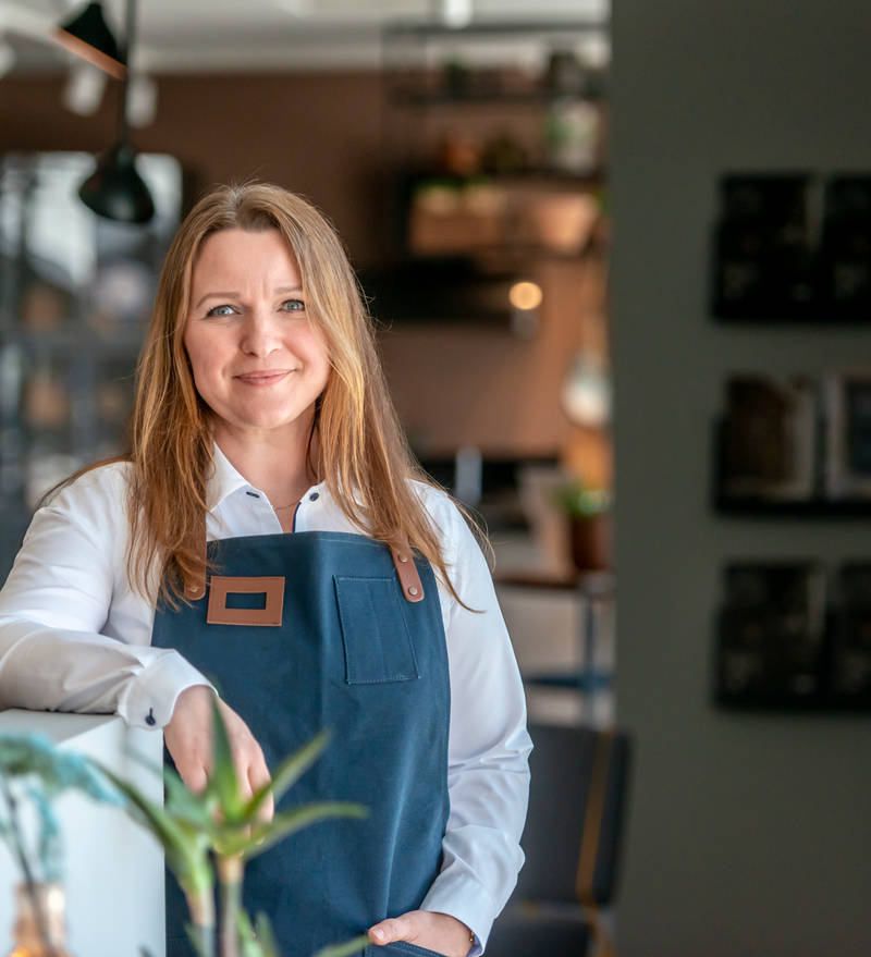 Kjøkkendesigner i lys grå skjorte og blått forkle smiler til kamera utenfor Sigdals butikk