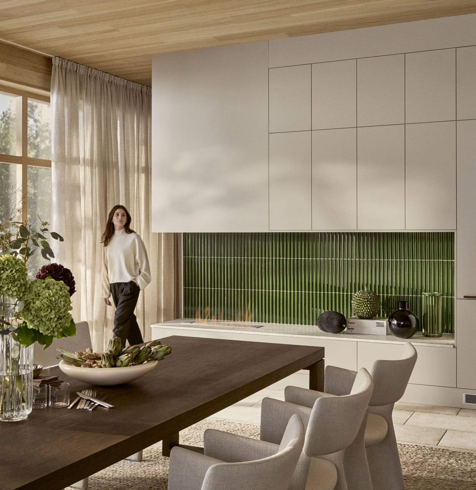 Kvinde kommer ind i rummet med spisebord i tre, benk og toppskabe Athena Sand med grønne fliser bak