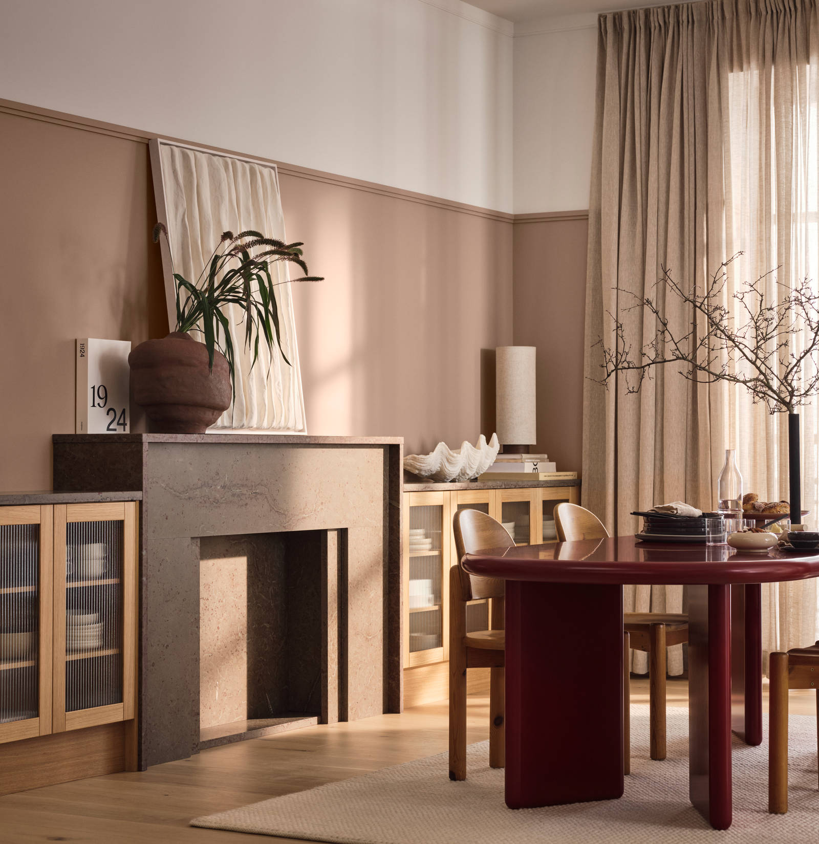 Öppen spis med växt i keramik-kruka intill vitrinskåp med träram i natur ek och räfflat glas, rött designat matbord