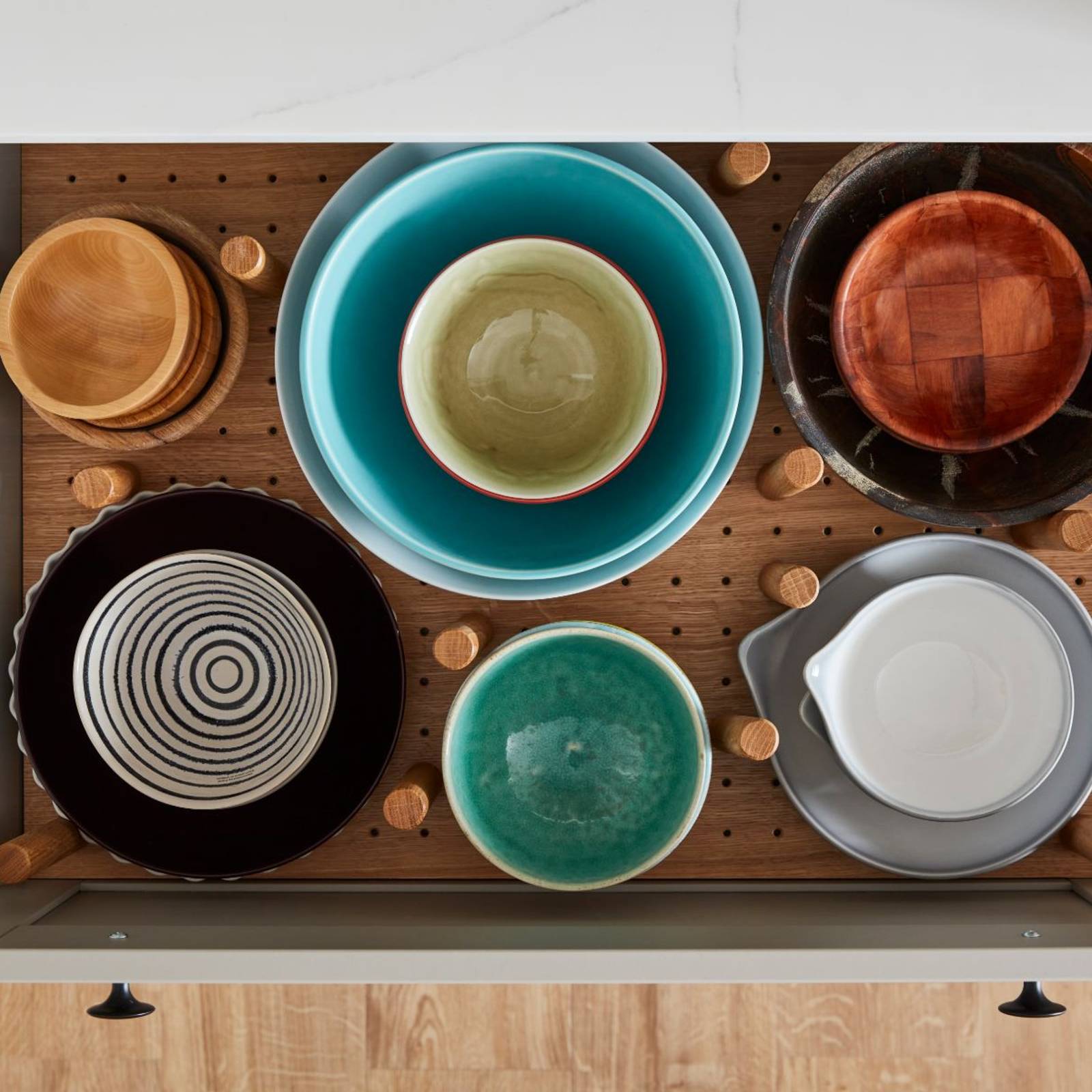 Opened drawer containing a storage solutions of large crockery organized by a peg board in the bottom of the drawer, with oak pegs holding them in place.