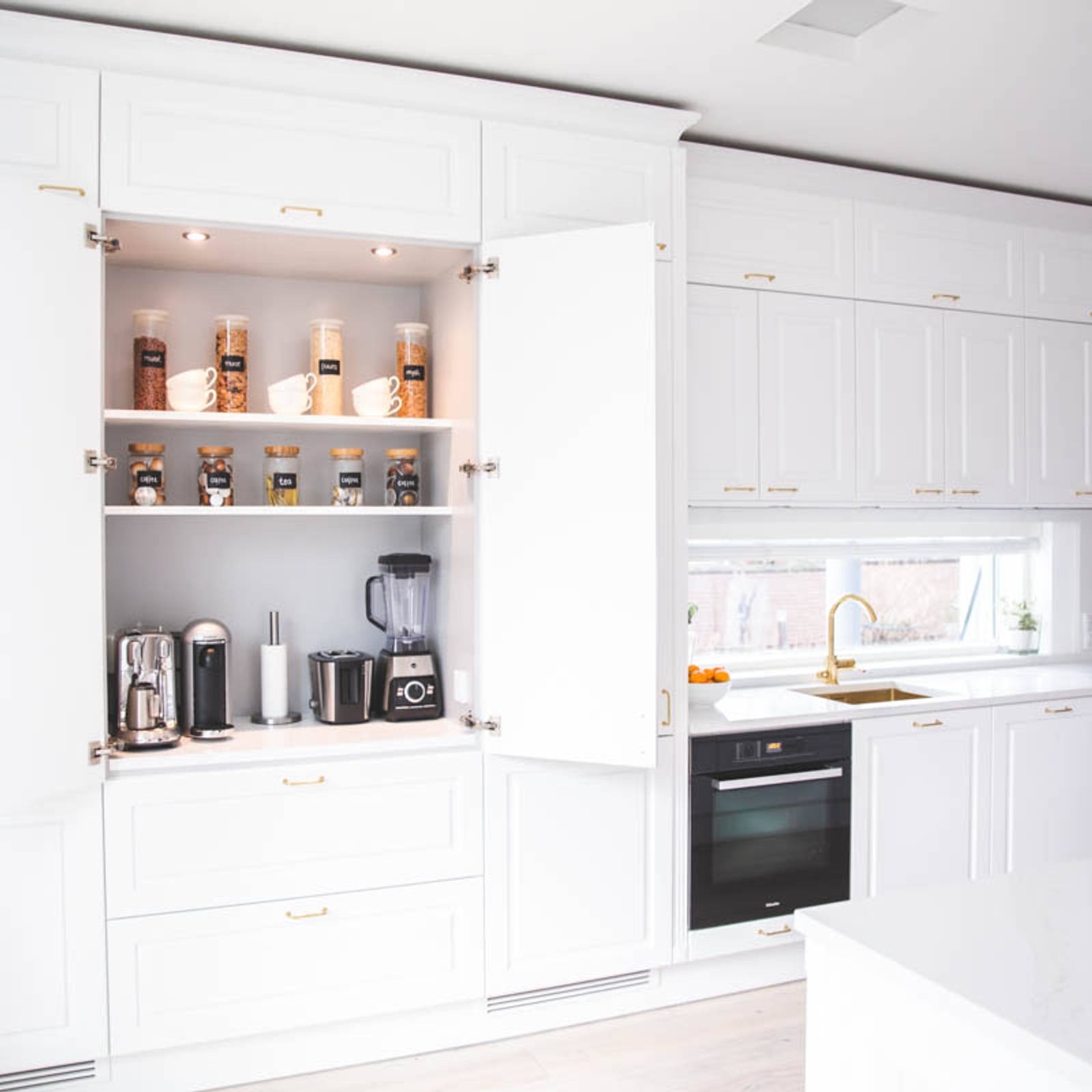 Valkoinen keittiö, jossa perinteiset maalatut karmiovet, messinkikahvat ja marmoroitu työtaso, avattu aamiaiskaappi kahvinkeittimillä, lasipurkit teetä ja kuppeja.